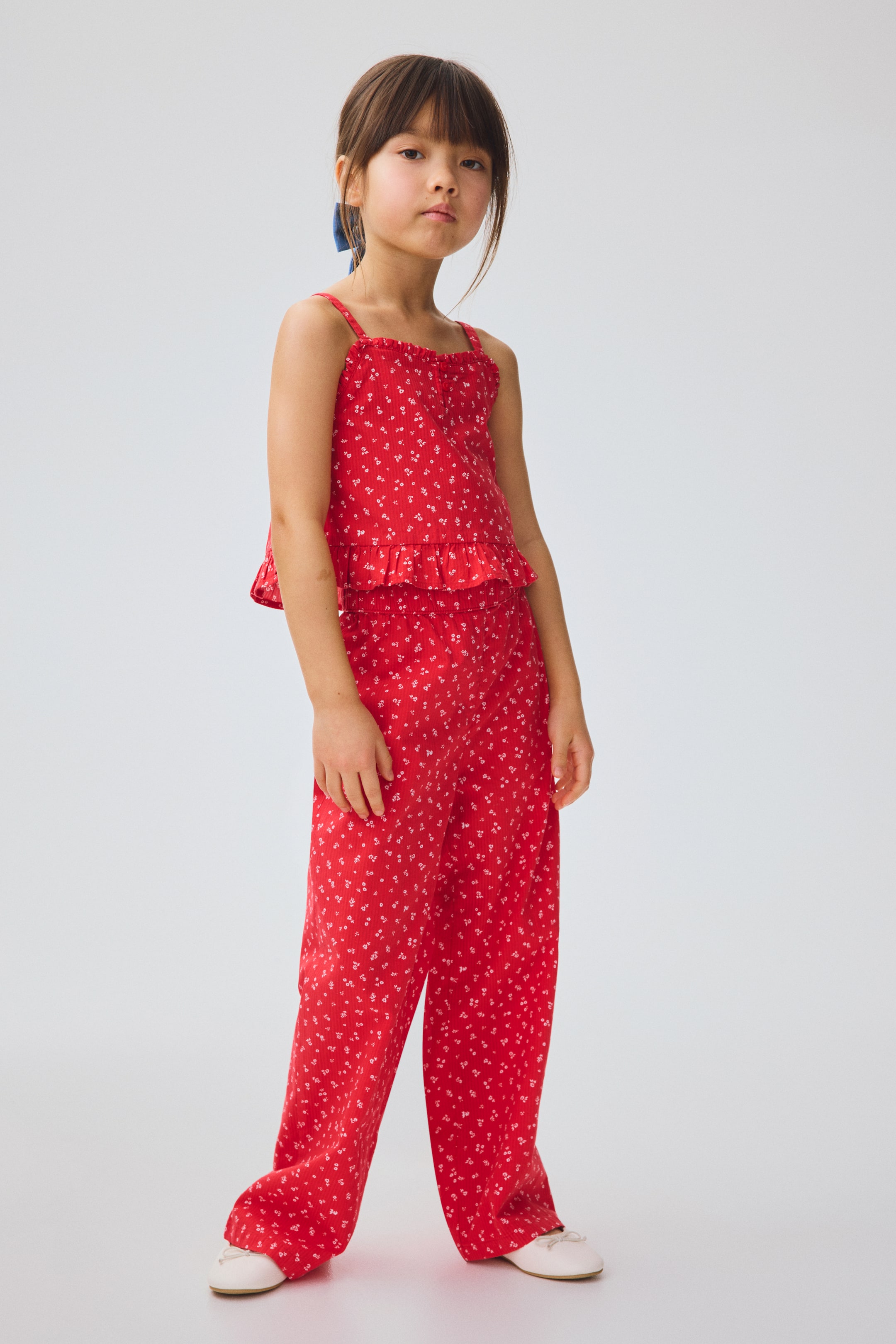 View larger image: A red two-piece set features a camisole top with thin spaghetti straps and a ruffled peplum hem, paired with matching wide-leg pants. The entire set showcases a small white floral print and a ribbed texture, presented from a slightly angled front view.
