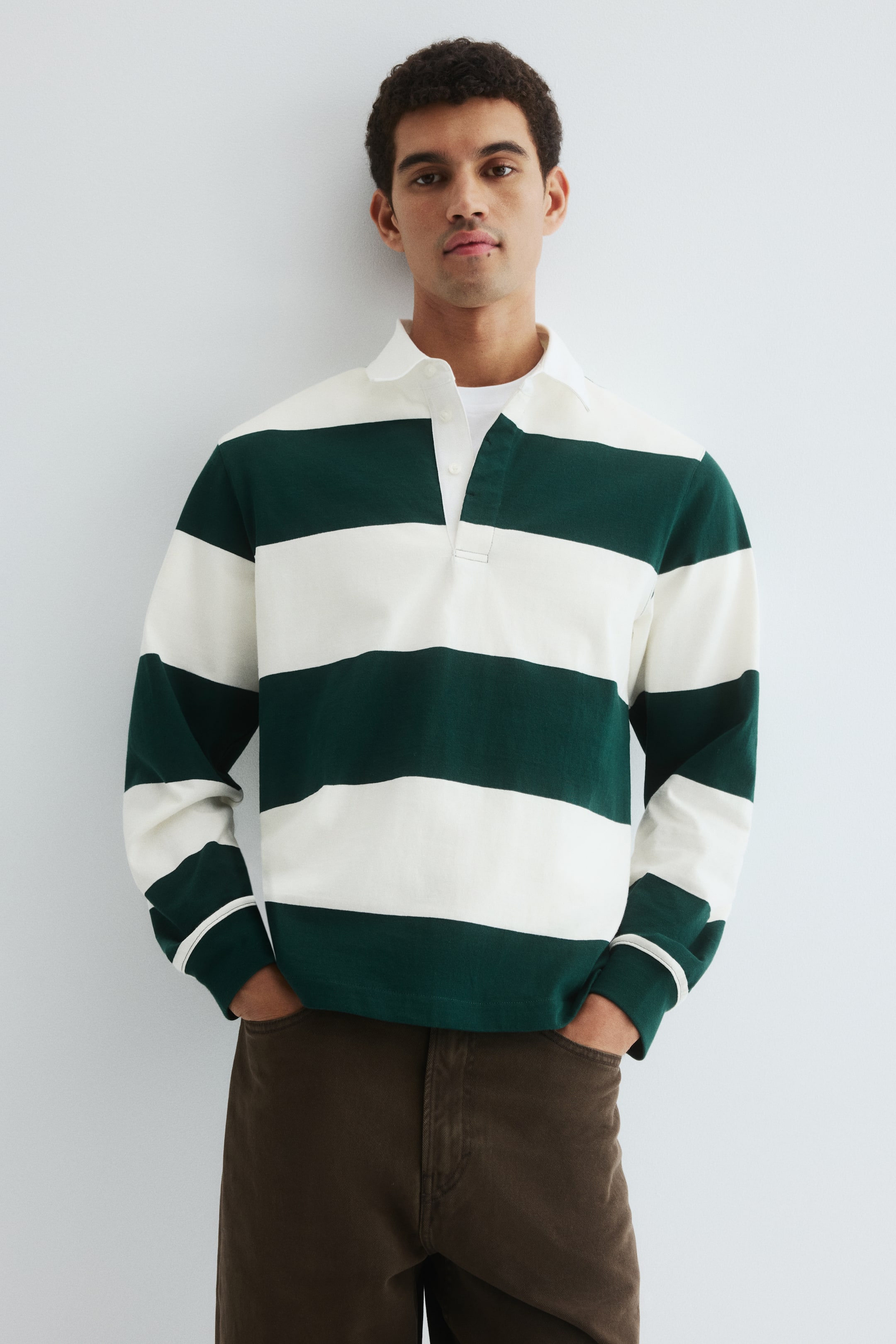 View larger image: A long-sleeved polo shirt is facing forward, displaying wide horizontal stripes in dark green and white. It is featuring a white polo collar and a three-button placket, worn open over a white t-shirt. The shirt’s long sleeves are ending in white ribbed cuffs, each detailed with two thin green stripes, and it is presenting a relaxed fit.