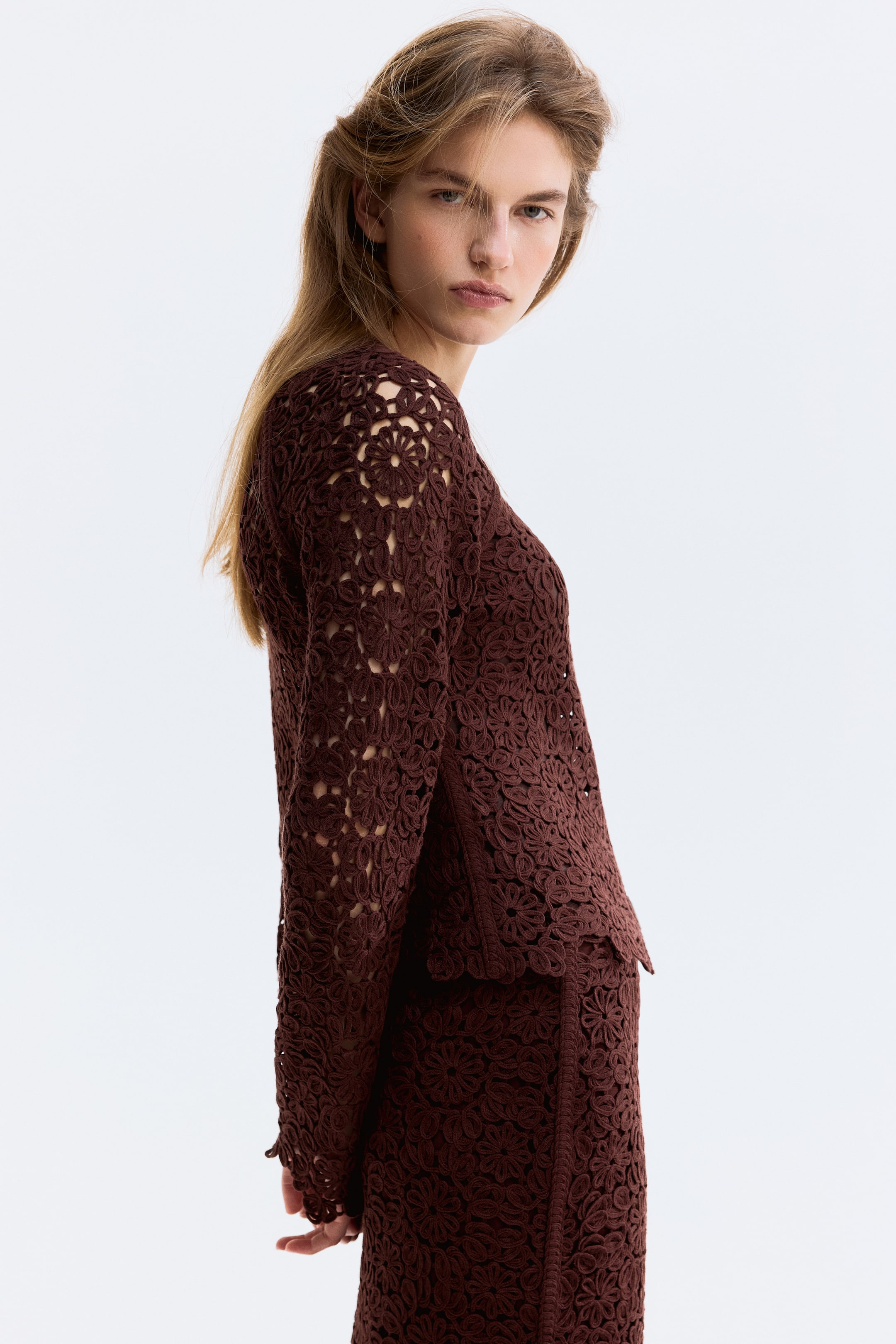 Ver imagen más grande: Un top de encaje floral marrón y una falda a juego exhiben un intrincado patrón floral calado. El top de manga larga y la falda son visibles desde un ángulo lateral-trasero, destacando su textura y forma.