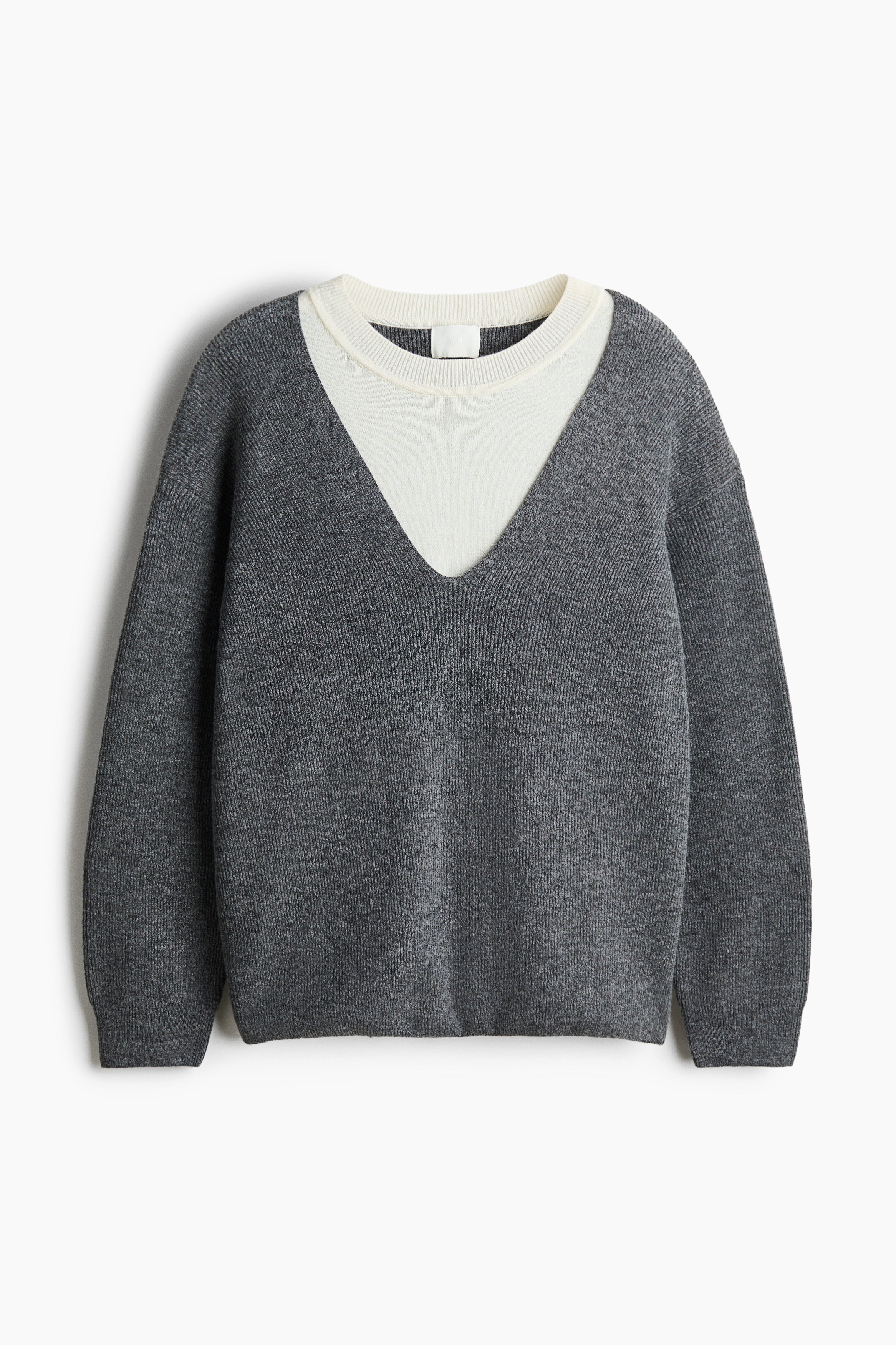 A layered knit jumper featuring a grey V-neck outer layer with dropped shoulders and long sleeves, and an integrated off-white inner layer with a round, rib-trimmed neckline, is worn by the model. The jumper is paired with blue corduroy trousers.