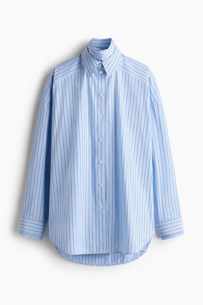 A light blue long-sleeved button-up shirt is displaying its front, featuring vertical stripes of alternating light blue and dark blue. It is designed with a pointed collar, a full button placket with white buttons, buttoned cuffs, and a curved hem, presenting an oversized fit. - 1