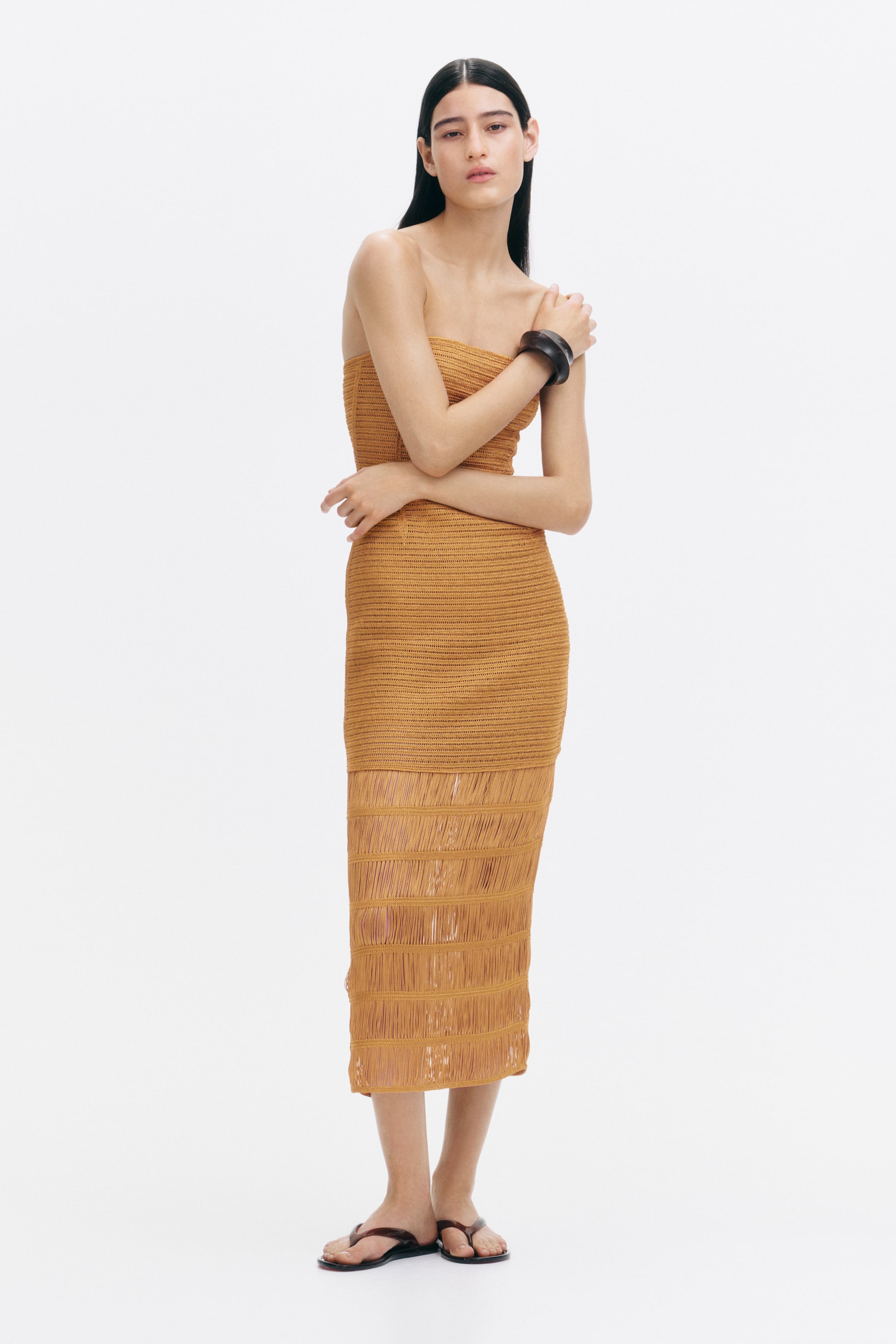Größeres Bild ansehen: Ein ockerfarbenes Bandeau-Kleid, gefertigt aus einem Strick mit Häkeloptik, verfügt über aufgenähte Fransenbesätze am Rock. Das Model trägt das Kleid, verschränkt die Arme und trägt zudem ein dunkles Armband und dunkle Flip-Flops.