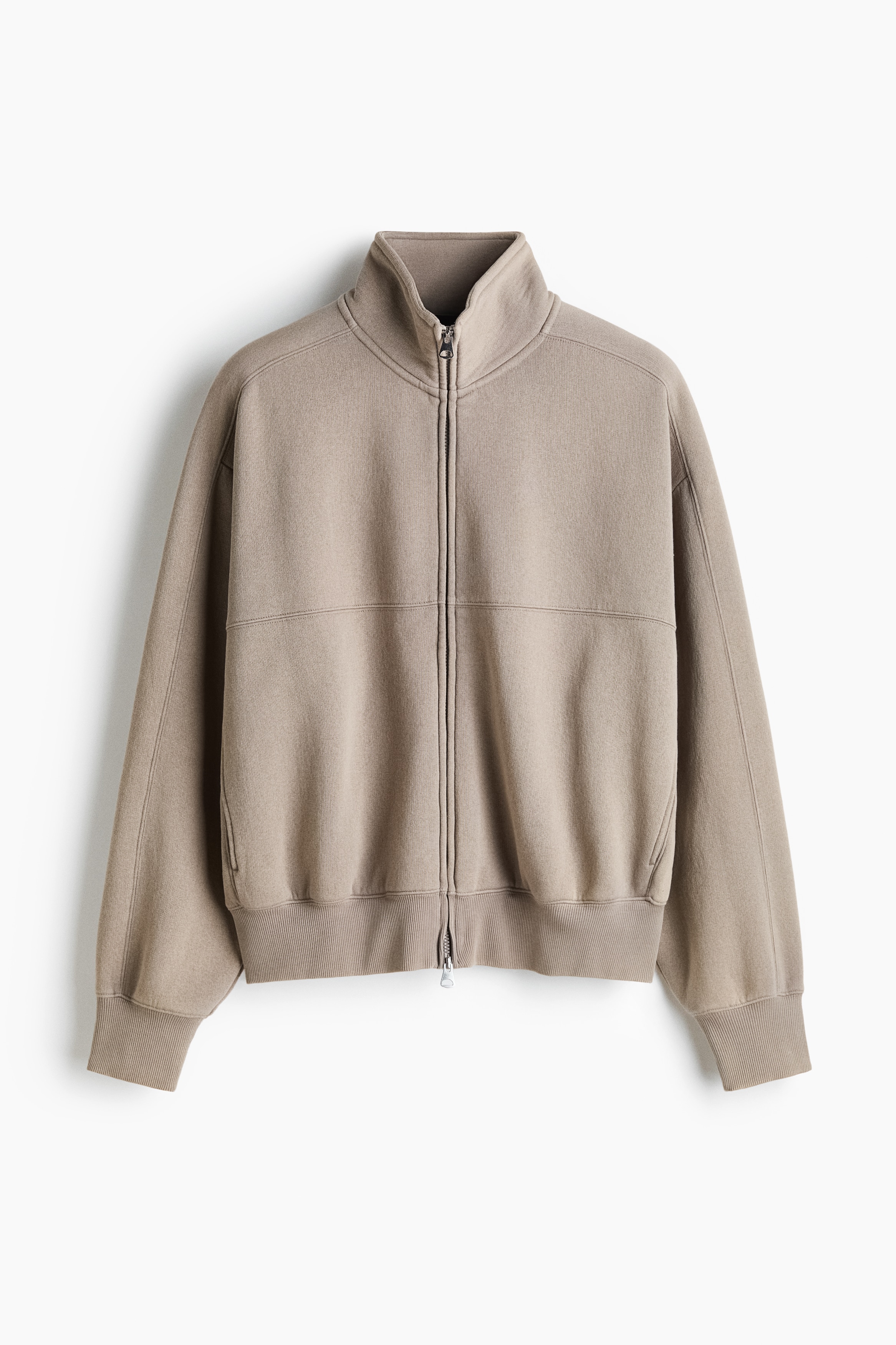 Une veste de survÃªtement beige Ã  coupe oversize est portÃ©e par le mannequin, qui a une main glissÃ©e dans une poche latÃ©rale discrÃ¨te. La veste prÃ©sente un col montant, une fermeture Ã©clair avant Ã  double sens et des finitions cÃ´telÃ©es aux poignets et Ã  lâourlet, agencÃ©e avec un pantalon assorti.