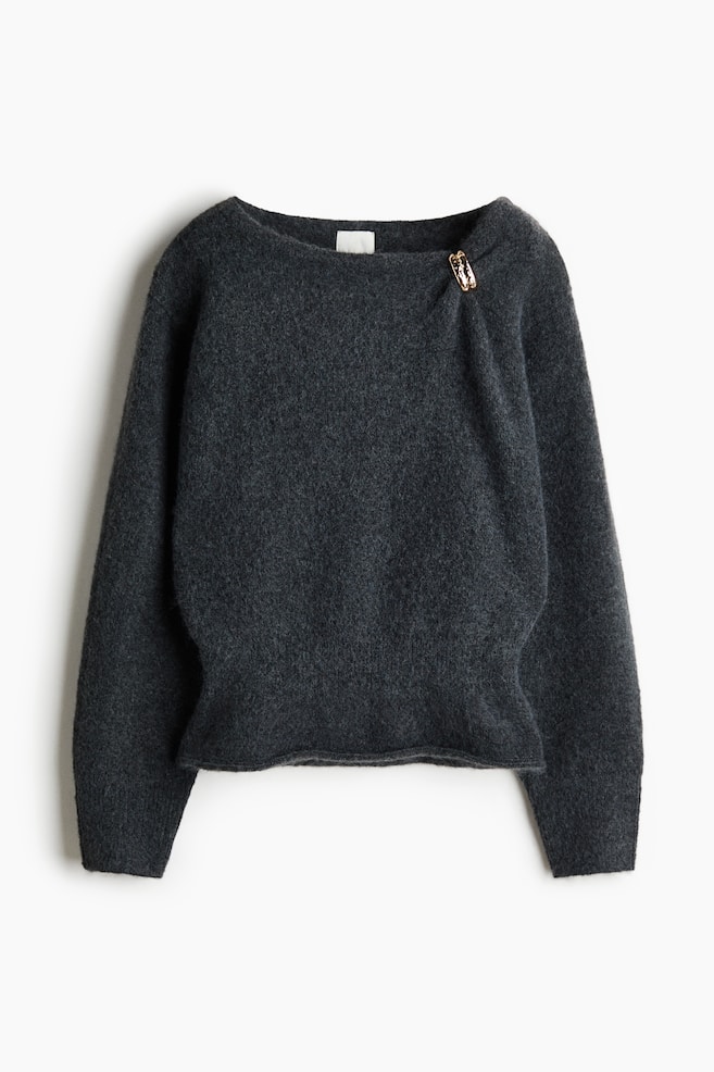 A dark grey brushed-knit jumper is shown from the front, featuring a wide neckline with an asymmetrical gather secured by a small gold-toned clasp on the left shoulder. The jumper has long sleeves and a fitted ribbed waistband that flares slightly at the hem. - 1