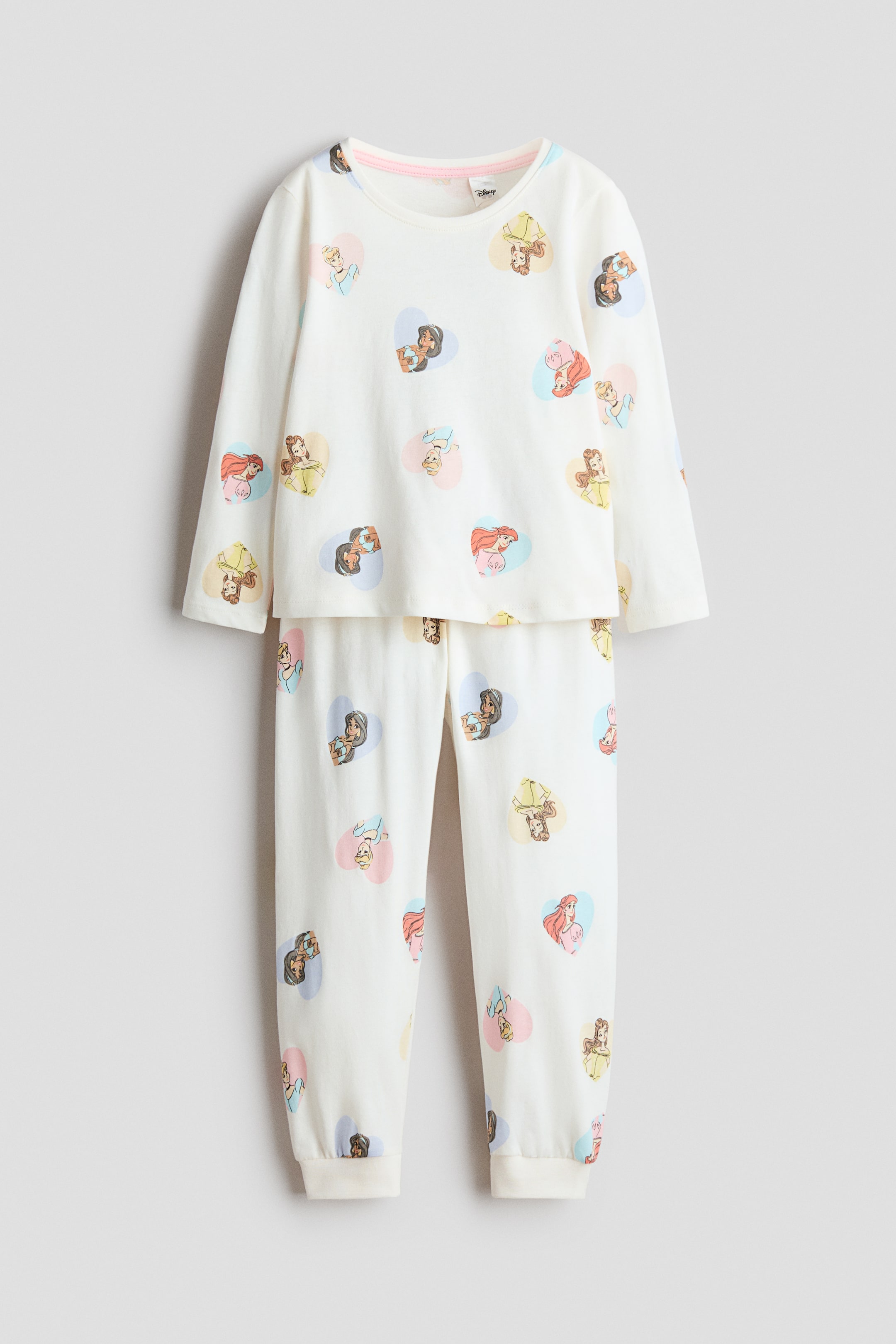 View larger image: A two-piece cream-colored pajama set is displaying, featuring a long-sleeved top with a round neckline and matching bottoms. Both pieces are showcasing a repeated pattern of various female cartoon characters inside pastel heart shapes, and the bottoms are finished with ribbed hems at the ankles.