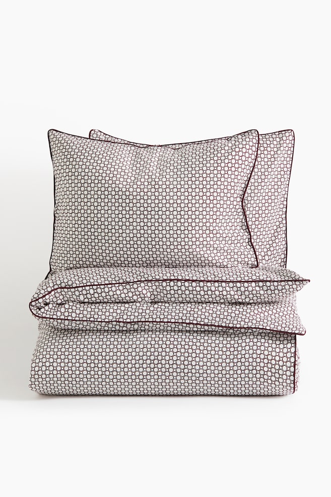 A bedding set consisting of a folded duvet cover and two upright pillowcases is facing front and displaying a repeating burgundy and white geometric grid pattern. Both the duvet cover and pillowcases feature thin burgundy piped edges along their borders. - 1