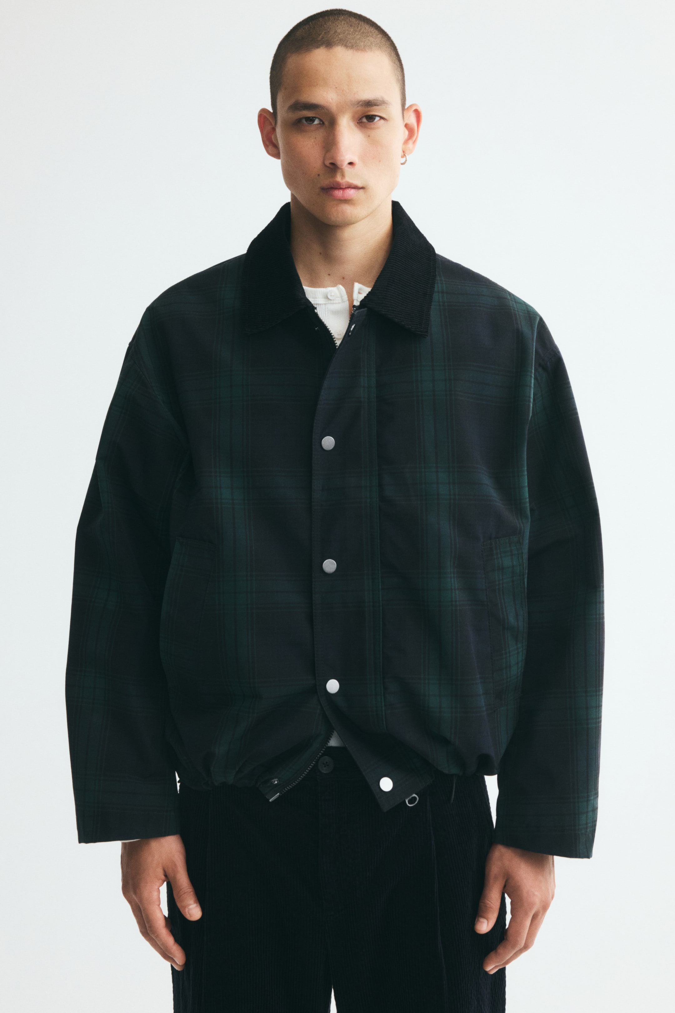 Agrandir l'image: Le mannequin porte une veste coach à carreaux vert foncé et noir par-dessus une chemise blanche et un pantalon noir. La veste présente un col en velours côtelé noir, des épaules tombantes, un ourlet resserré élastiqué et un rabat coupe-vent frontal avec des boutons-pression argentés.