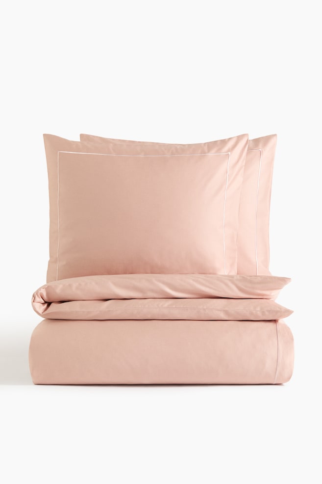 Un ensemble de literie rose poudré comprend deux taies d'oreiller orientées vers l'avant avec des bordures cousues blanches, soigneusement empilées au-dessus d'une housse de couette pliée affichant un détail de bordure assorti. - 1