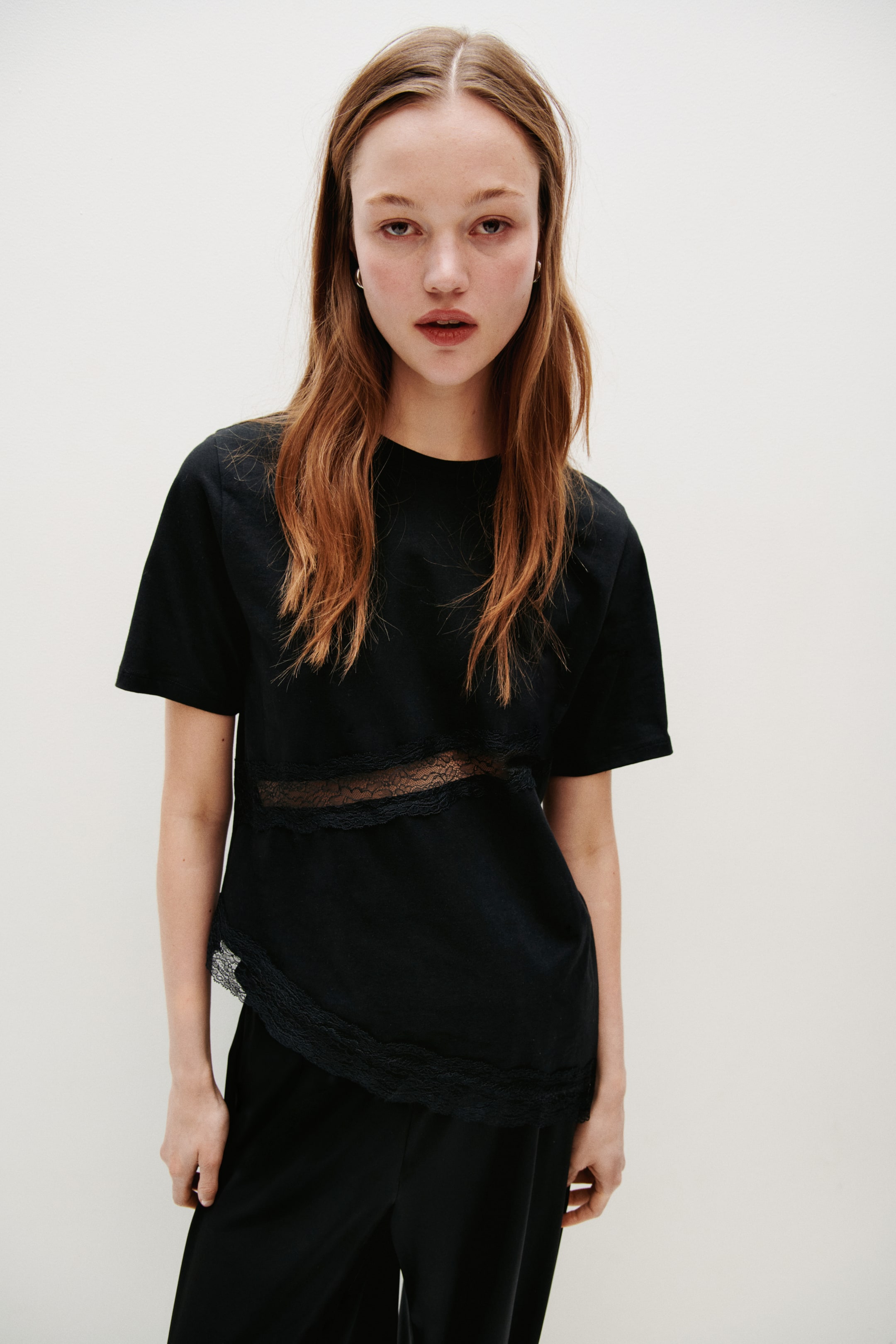 Vis større bilde: En svart kortermet topp vender mot betrakteren, med et gjennomsiktig horisontalt blondedetalj over midtpartiet. Den har også blondekanter langs topp- og bunnkanten av panelet, samt langs den asymmetriske nedre venstre kanten.