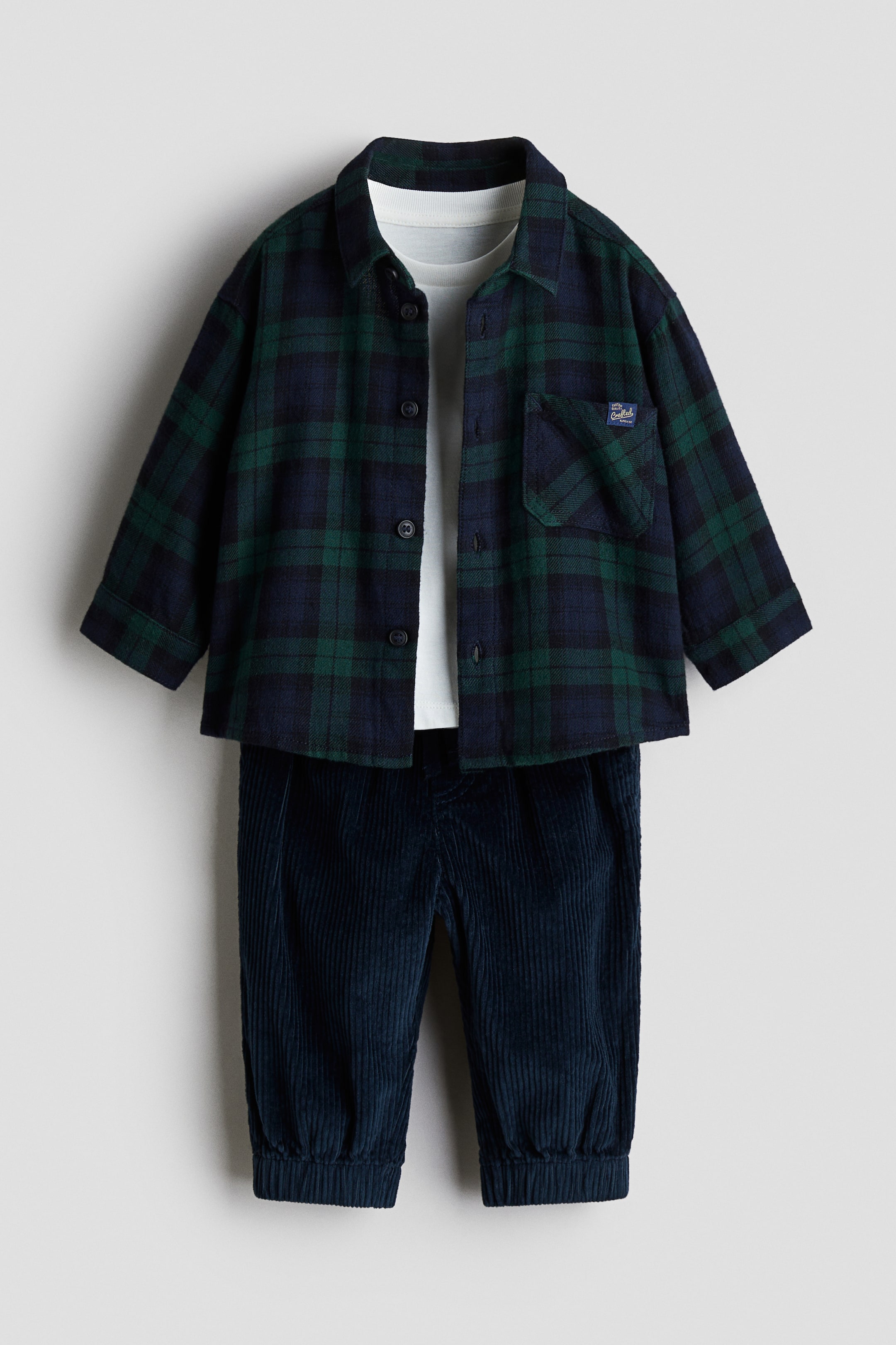 Voir une image plus grande: Un ensemble en coton de 3 pièces comprend une chemise à carreaux verts et bleu marine boutonnée sur le devant, qui présente une poche poitrine et est ouverte, révélant un t-shirt crème à col rond en dessous. Un pantalon en velours côtelé bleu marine foncé, montrant une texture côtelée et des poignets élastiqués, complète l'ensemble.