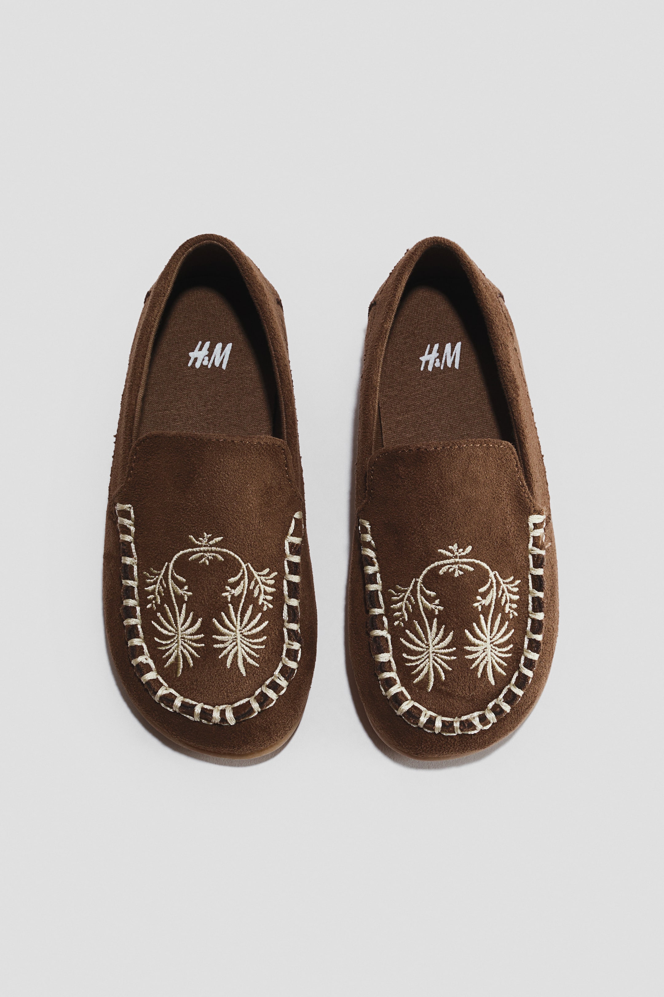 View larger image: Two brown moccasins, made from a suede-like material, are presented side by side, facing forwards. Each moccasin features light-coloured lacing around the toe box and an embroidered botanical pattern on the vamp. The H&M logo is visible in white on the brown inner sole.