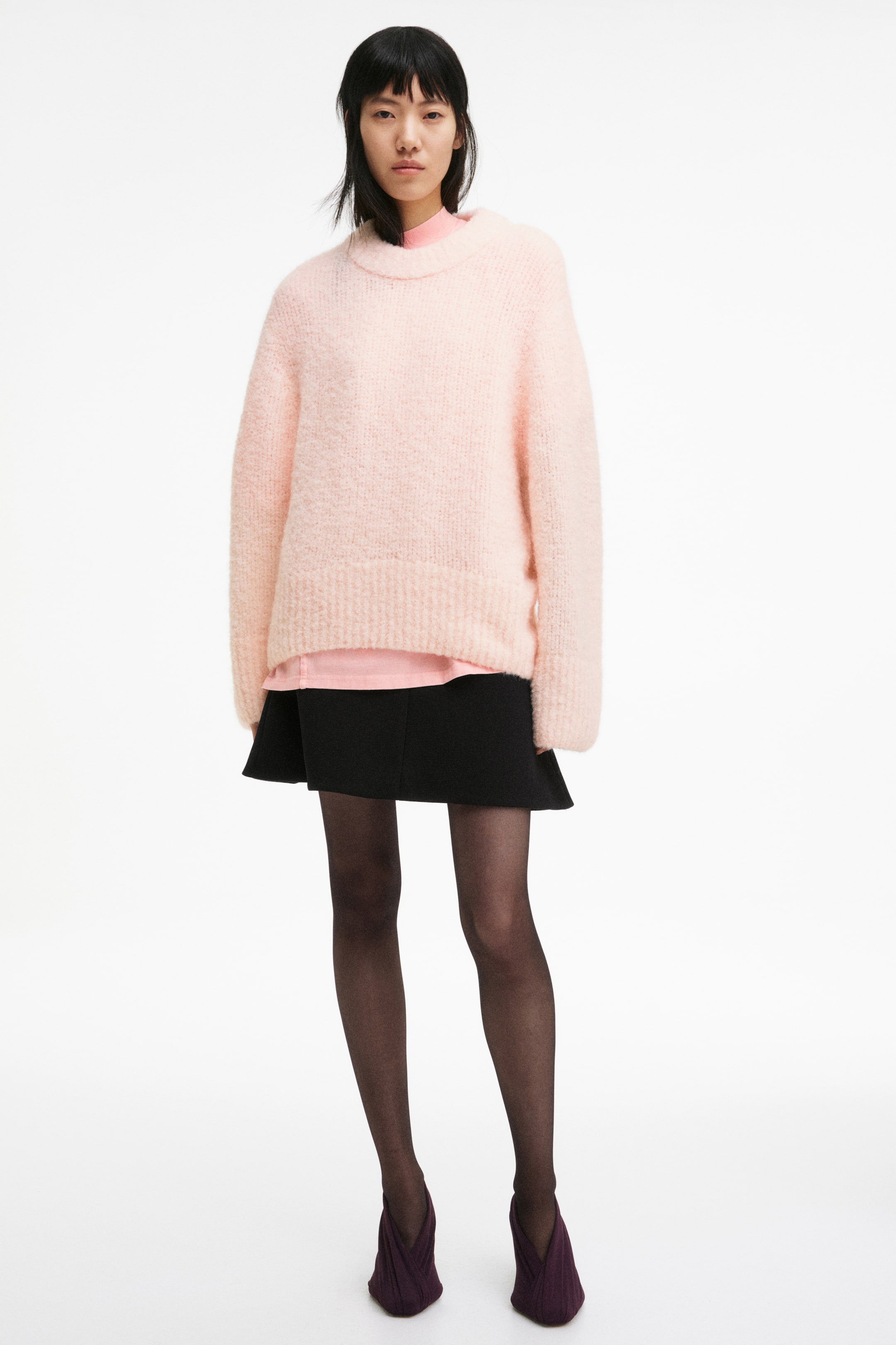 View larger image: The model is wearing a light pink, loose-fit jumper with a soft, woolly knit texture. It features a chunky, ribbed crew neck, dropped shoulders, and wide ribbing at the cuffs and hem, layered over a lighter pink top.