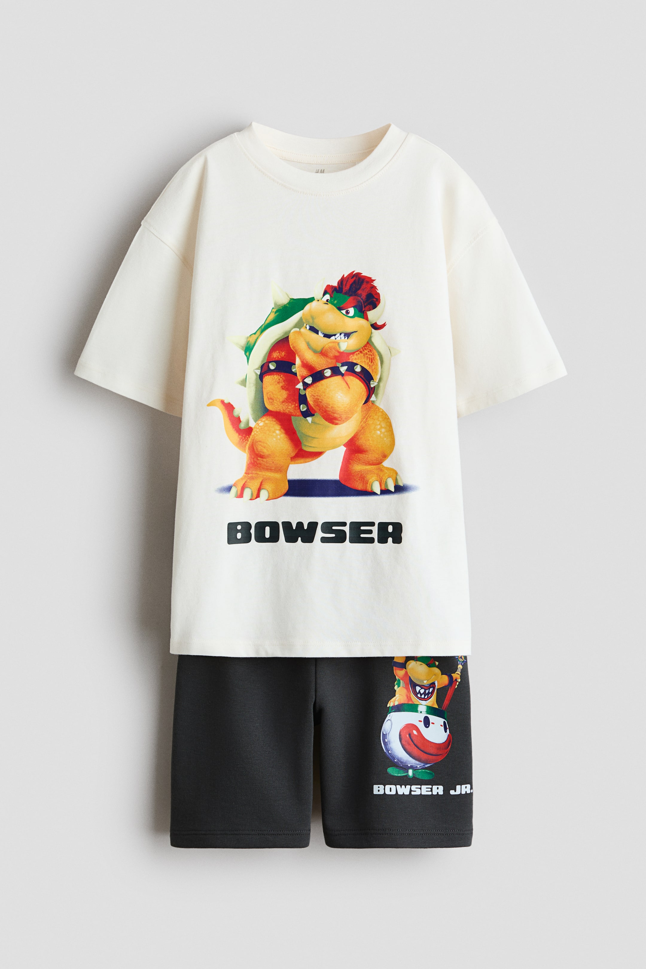 Vezi imaginea mai mare: Un set asortat este format dintr-un tricou din jerseu de bumbac alb murdar și pantaloni scurți din sweatshirt ușor, de culoare gri închis, ambele orientate spre față. Tricoul prezintă umeri căzuți, mâneci largi, o bandă de rib la gât și un imprimeu mare cu Bowser și cuvântul „BOWSER” dedesubt, în timp ce pantalonii scurți au elastic acoperit în talie și un imprimeu cu Bowser Jr. în mașina sa de clovn, cu „BOWSER JR.” dedesubt, pe piciorul drept.