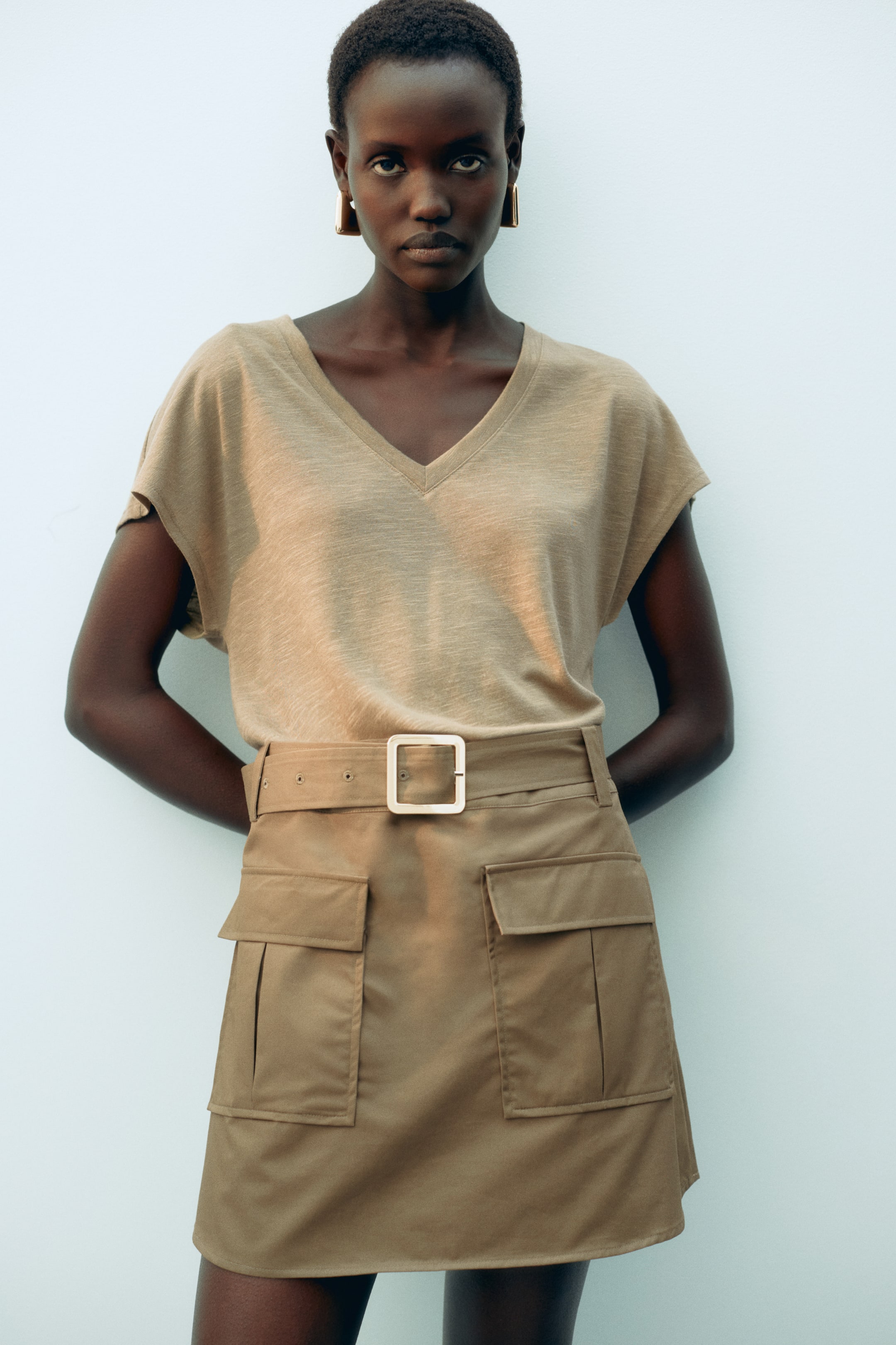 View larger image: A light-brown V-neck cap-sleeved top is tucked into a beige mini-skirt, which features a belted waist with a square buckle and two large flap utility pockets on the front.
