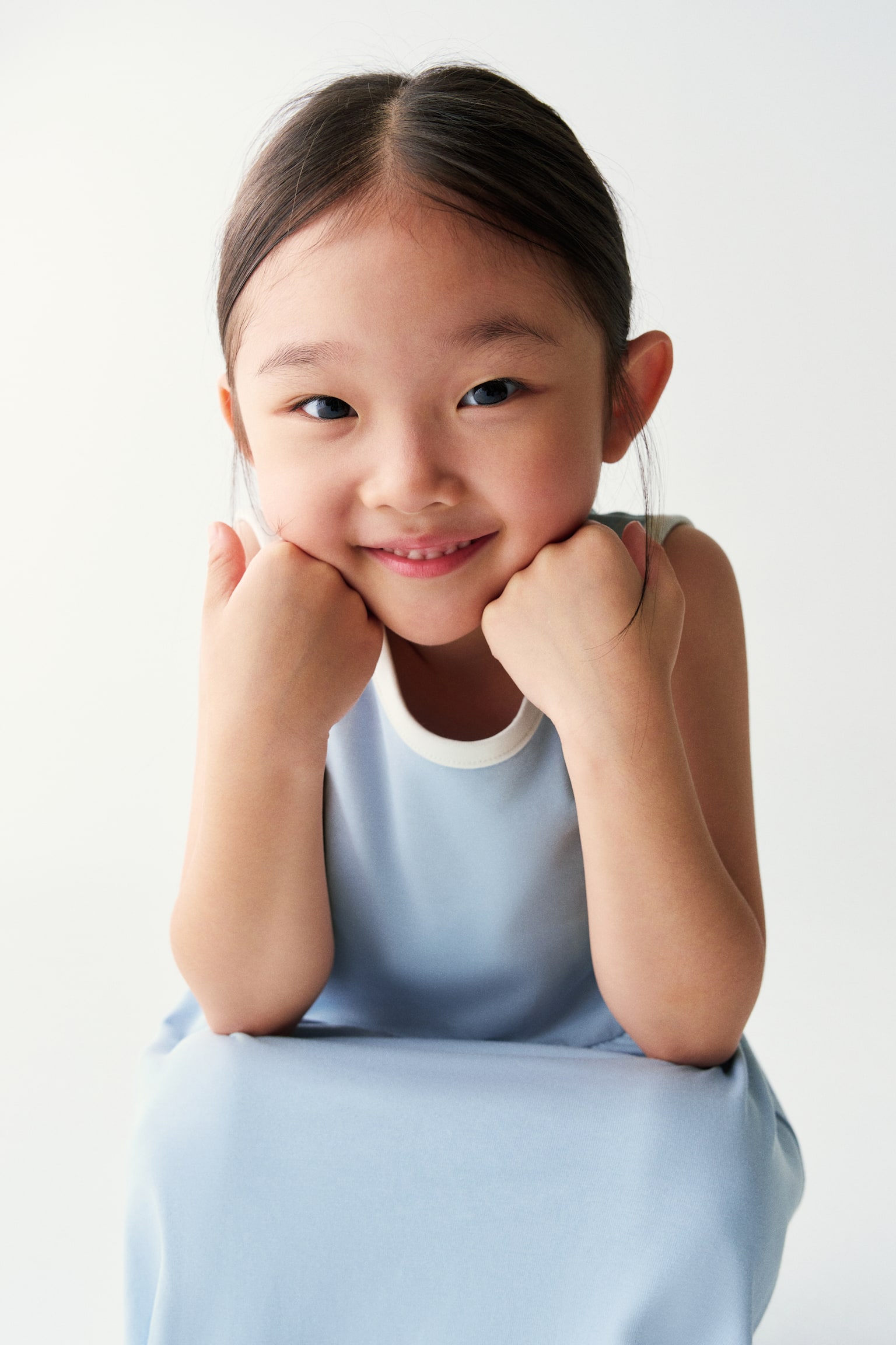 A light blue sleeveless dress with contrasting white trim at the round neckline is worn by the model. She is posing with her chin resting on her hands, highlighting the garmentâs clean, simple design. - 1