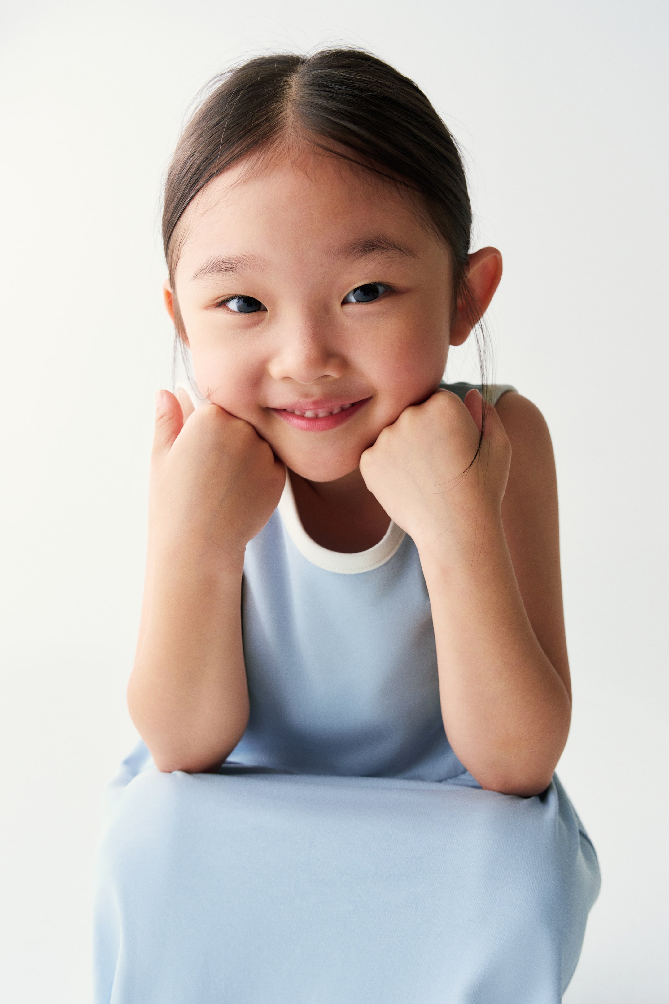 View larger image: A light blue sleeveless dress with contrasting white trim at the round neckline is worn by the model. She is posing with her chin resting on her hands, highlighting the garment’s clean, simple design.