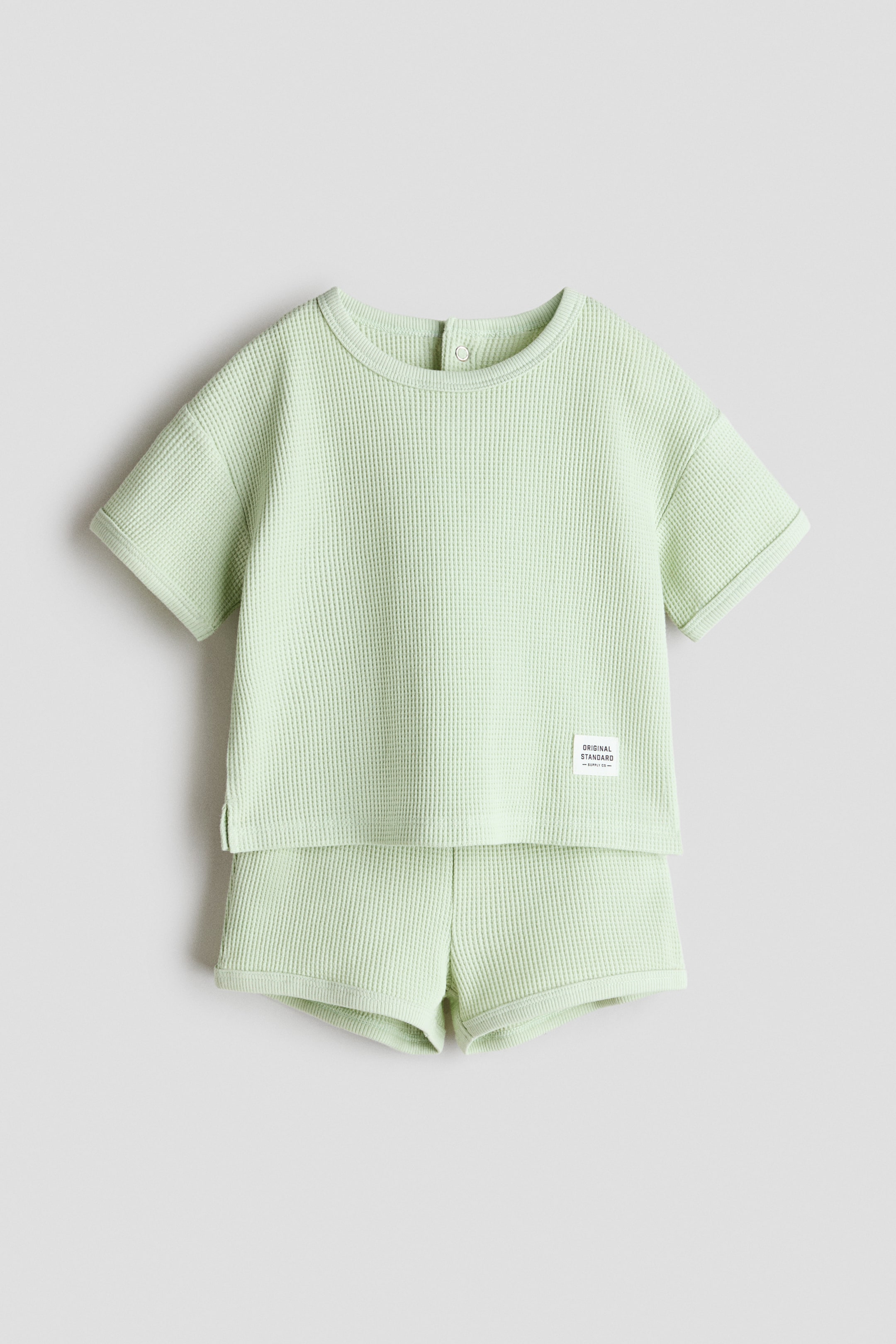 View larger image: A light green waffled jersey set is shown from the front, highlighting a t-shirt with a round, ribbed neckline, a visible press-stud closure at the back of the neck, ribbed short sleeve cuffs, and a small white label on the lower right front. The matching shorts feature a plain waistline and waffled texture.