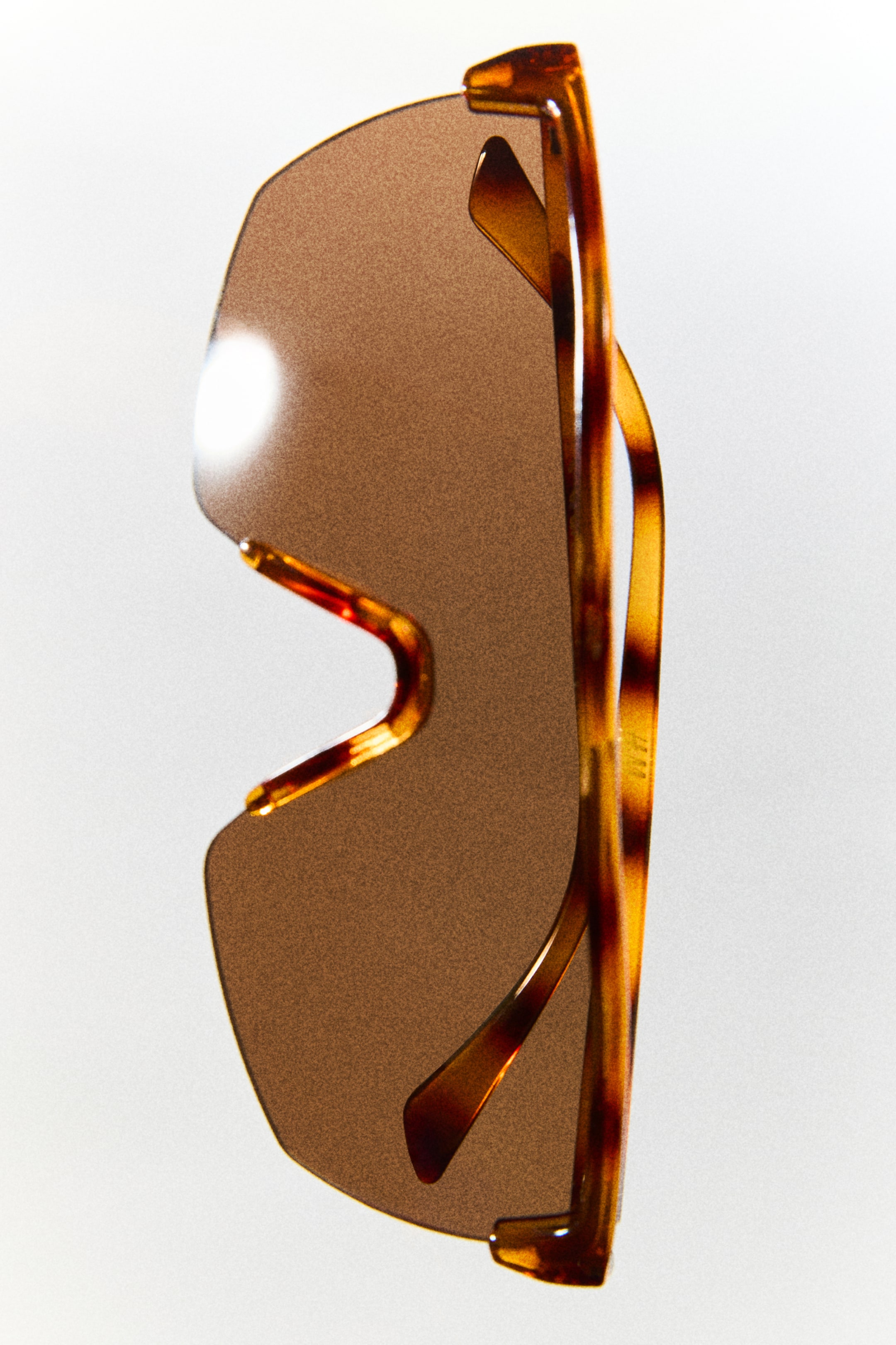 View larger image: Shield sunglasses featuring a single, continuous brown-tinted lens and a tortoiseshell-patterned plastic frame. The sunglasses are positioned vertically, showing the mottled brown and amber temples folded behind the lens.