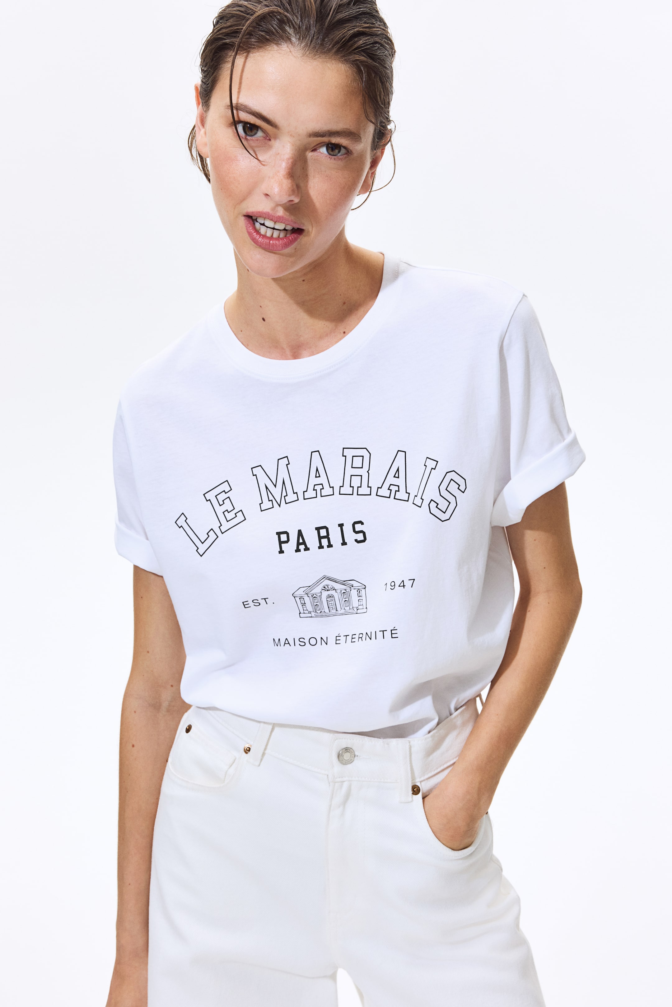 View larger image: A white t-shirt features a black graphic print on the front, showcasing "LE MARAIS" in arched outline letters, "PARIS" in solid letters, and an illustration of a building facade with "EST. 1947" and "MAISON �TERNIT�". It is crafted with a round neckline and short sleeves that are rolled up.