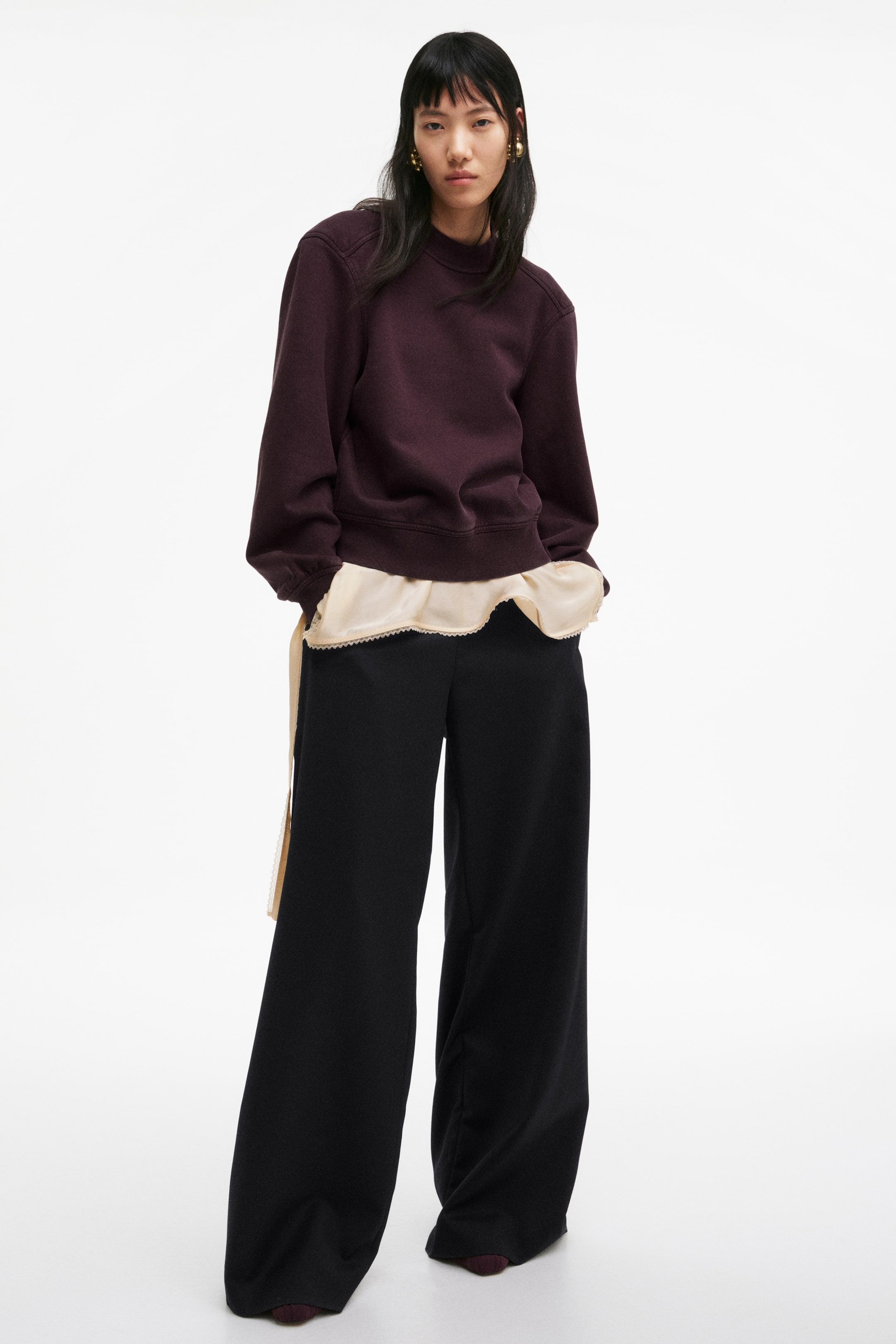 View larger image: A dark purple crewneck jumper features a relaxed fit with long sleeves, ribbed cuffs, and a ribbed hem, slightly angled towards the viewer's right. It is worn over a light-coloured, ruffled top that peeks out below its hem.
