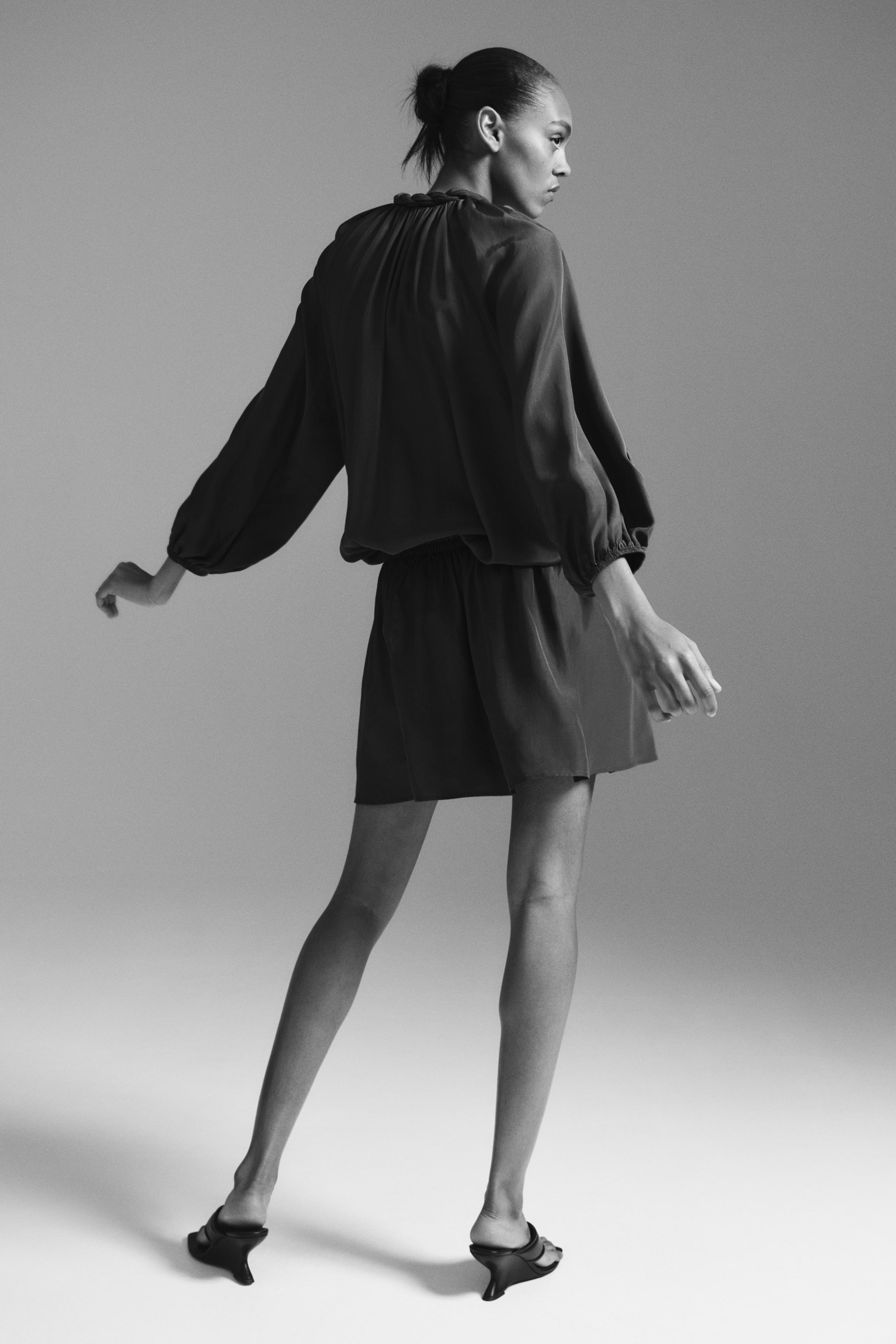 Nagyobb kép megtekintése: A romper egy magas, ráncolt nyakkivágással és hosszú, ráncolt mandzsettás ballon ujjakkal rendelkezik, hátulról nézve. A felső rész blúzosan omlik az elasztikus derék fölé, és a laza szabású rövidnadrág térd fölé ér.