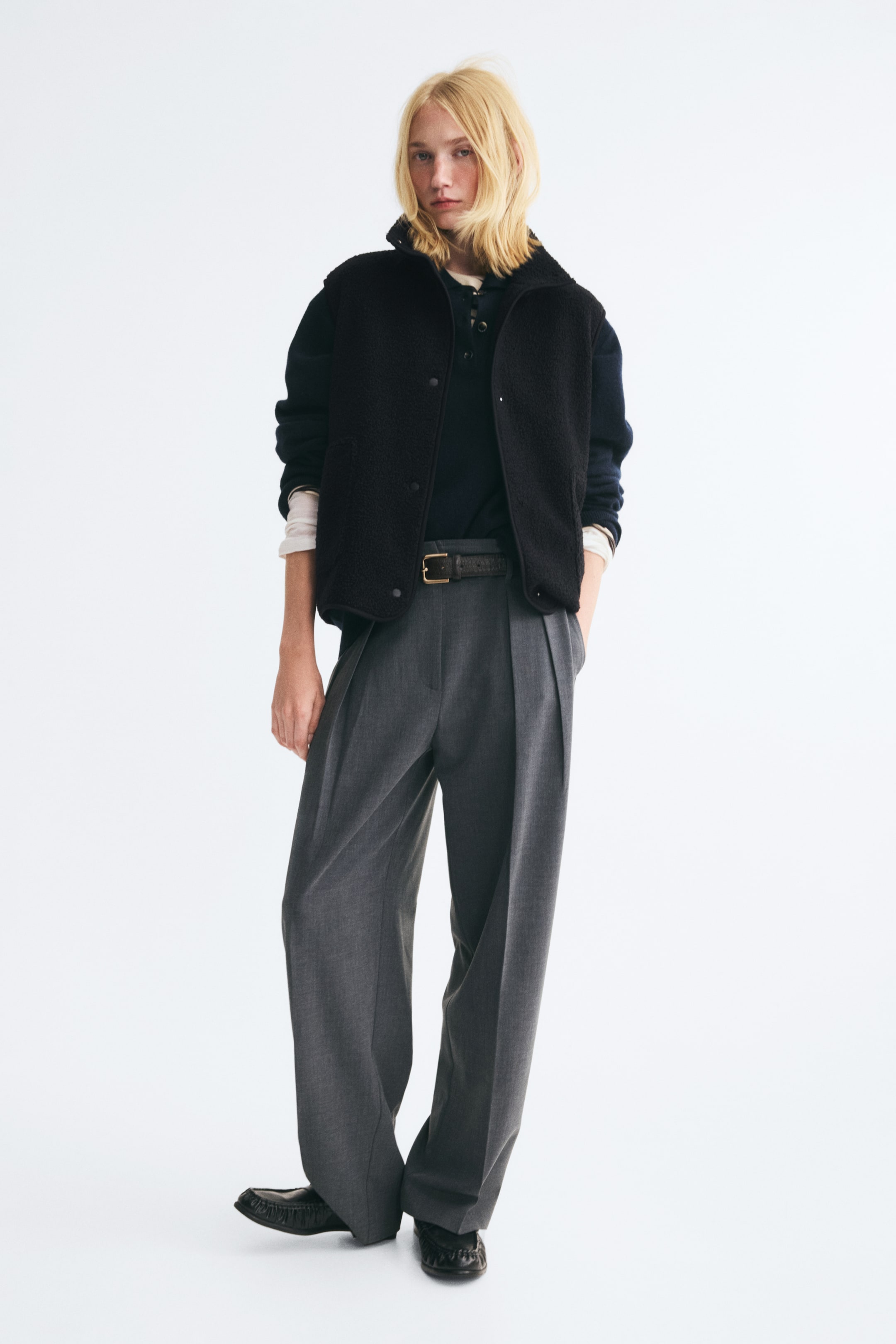 View larger image: A black teddy vest featuring a funnel collar and front press-stud fasteners is worn open by the model. The vest is layered over a dark long-sleeved top and styled with grey wide-leg trousers.