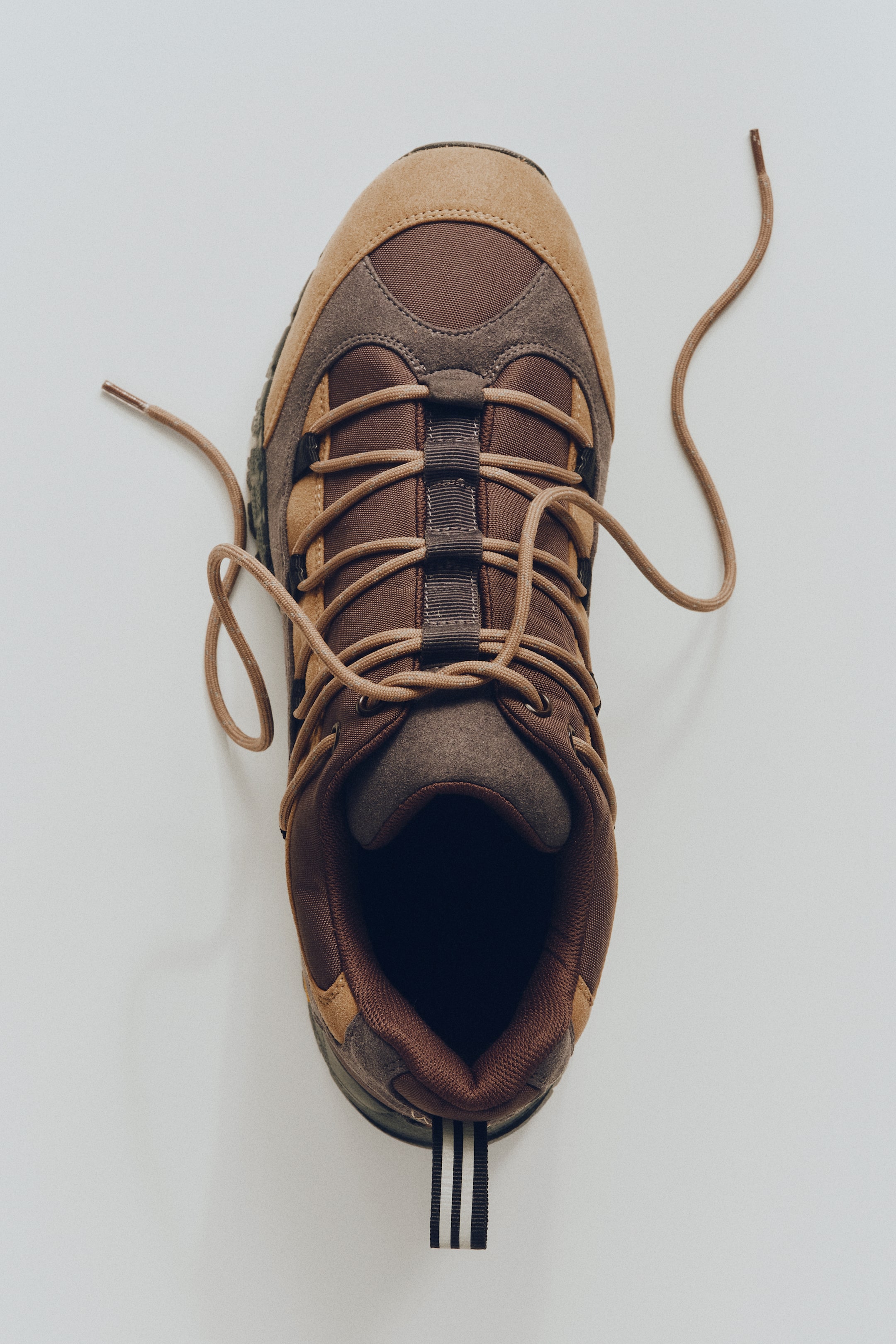 Visualizza immagine più grande: Una singola scarpa da trekking è vista direttamente dall'alto. Presenta pannelli in camoscio color beige e tessuto marrone scuro, con lacci marrone chiaro che passano attraverso anelli di tessuto e cadono morbidi, e un passante nero con tre strisce bianche è visibile sul tallone.