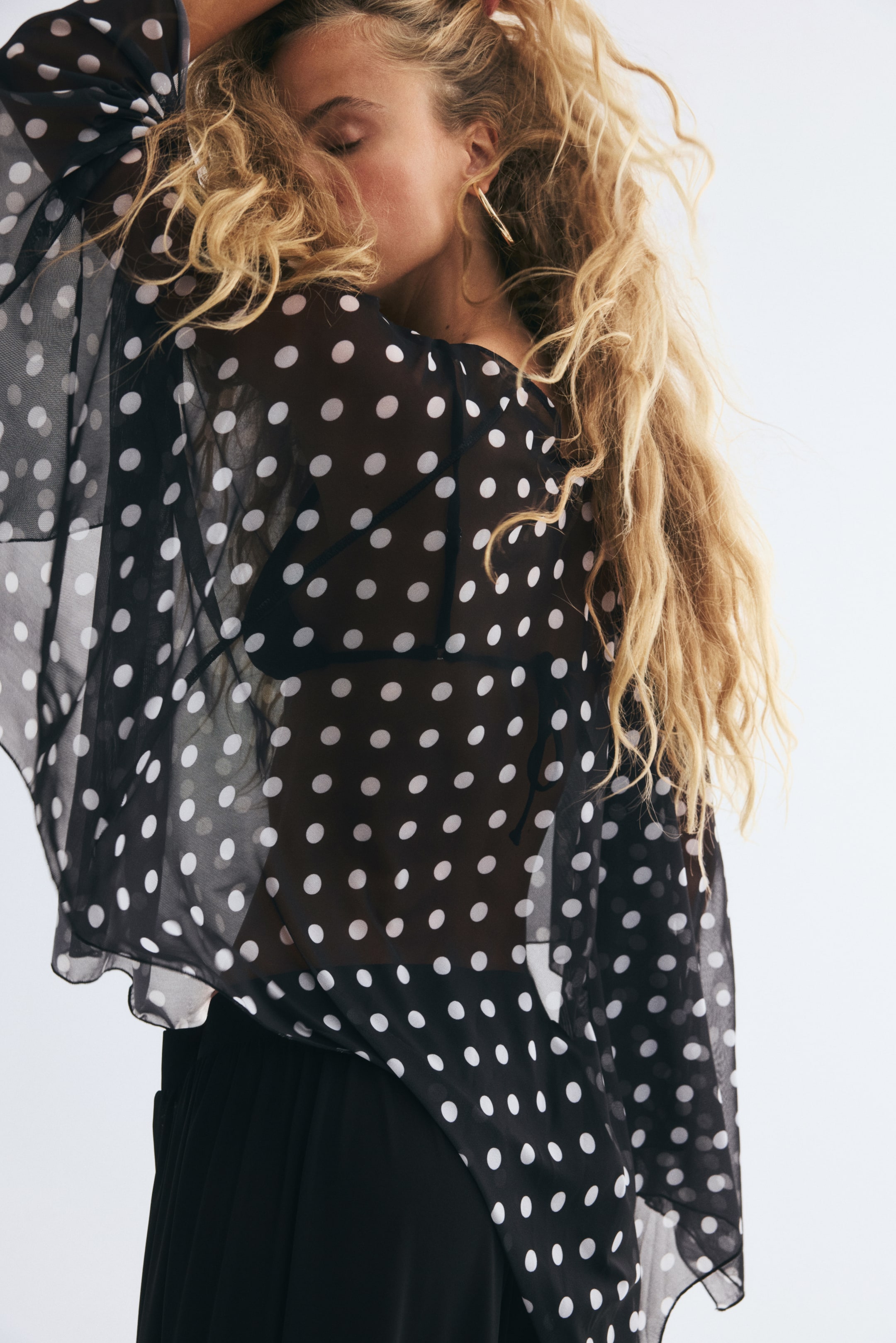 Agrandir l'image: Une cape en maille transparente à motif à pois noirs et blancs est portée par le mannequin. Le vêtement présente une silhouette drapée avec un ourlet asymétrique et est superposé sur un haut et un bas noirs.