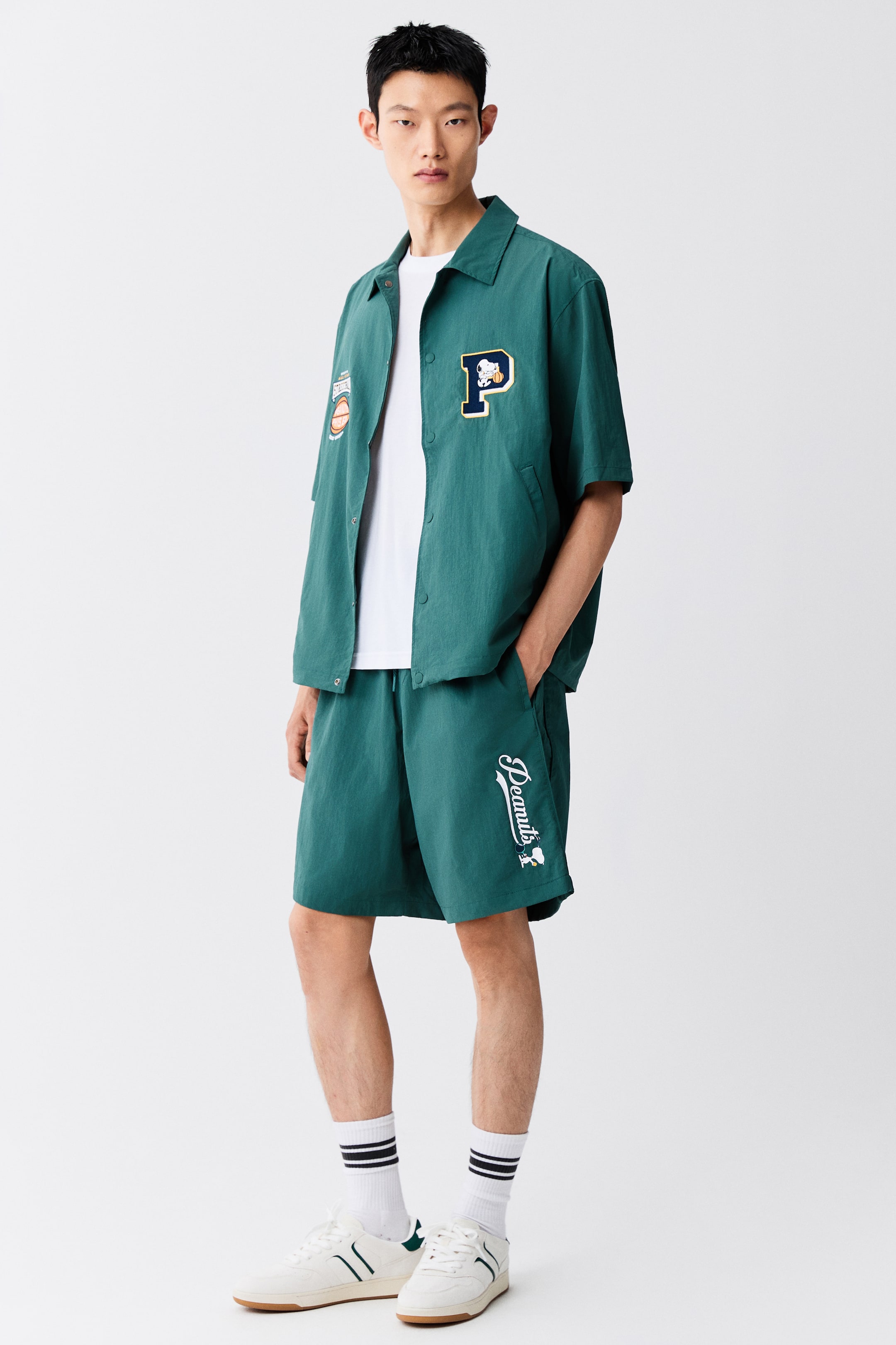 View larger image: Green knee-length track shorts with a relaxed fit and a white script graphic on the left leg are worn by the model, with one hand resting in the side pocket. The shorts are styled as a set with a matching green short-sleeved button-up shirt layered over a white T-shirt.