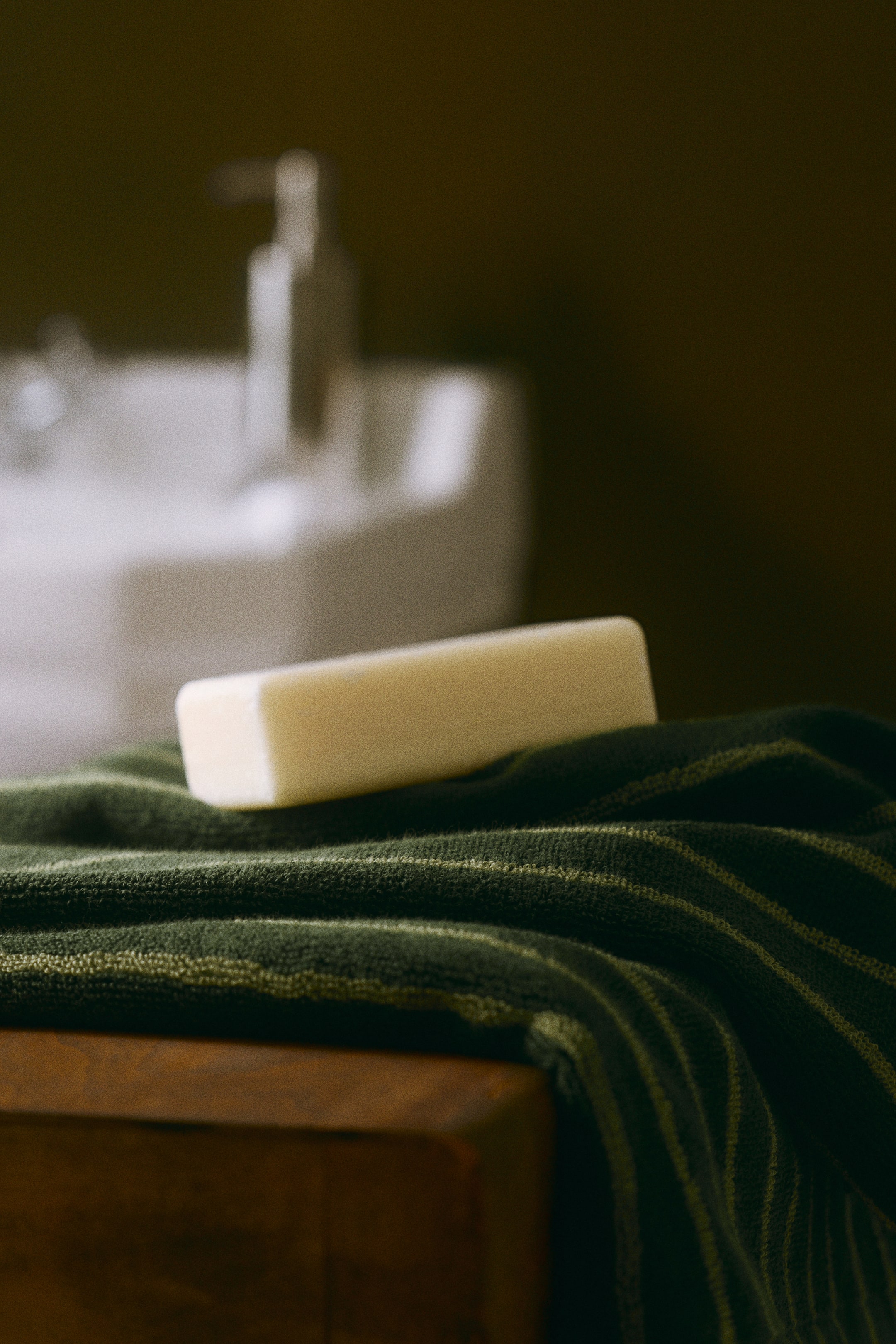 Ampliar la imagen: Una toalla de mano de felpa de algodón verde oscuro con delgadas franjas verde claro cuelga sobre el borde de madera, con una barra rectangular de jabón color crema descansando sobre su superficie doblada.