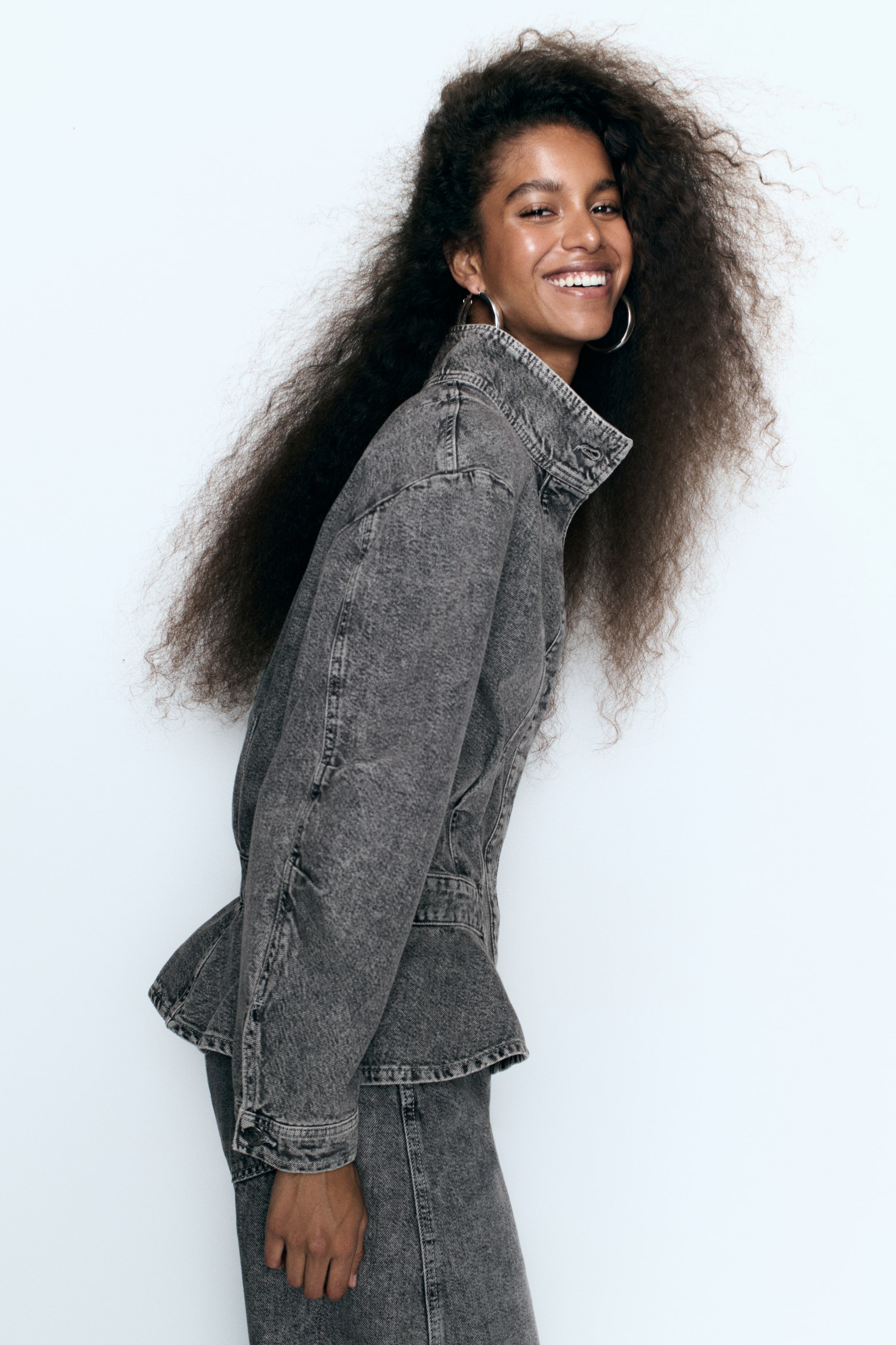 Vezi imaginea mai mare: Un model poartă o cămașă din denim gri, cu guler tunică, umeri căzuți și tiv peplum evazat. Cămașa este asortată cu pantaloni din denim gri, iar imaginea este prezentată din profil lateral.