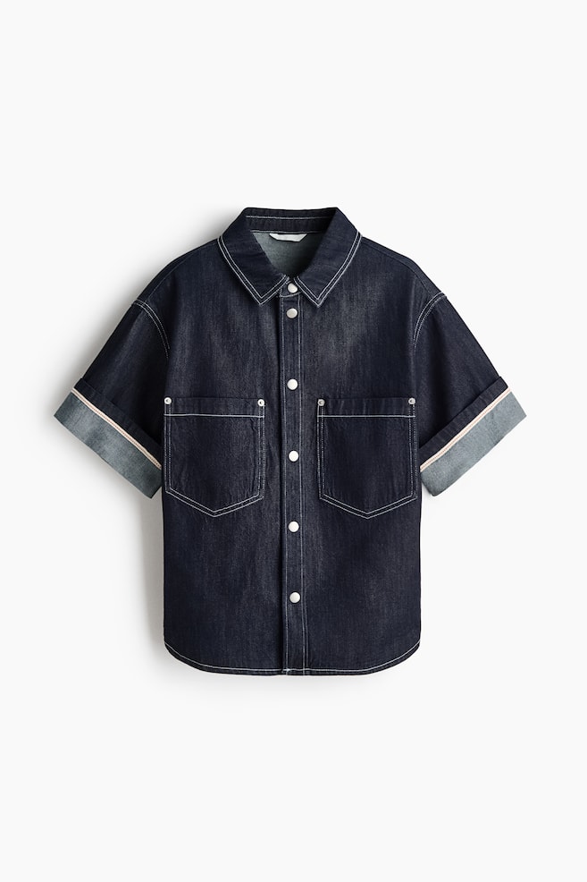 A relaxed-fit shirt in dark blue cotton denim is showcased from the front, highlighting a collar, press-studs along the front, and striking white contrast stitching throughout. It features two patch chest pockets, dropped shoulders, and short sleeves with turn-up cuffs revealing a red and white striped detail. - 1
