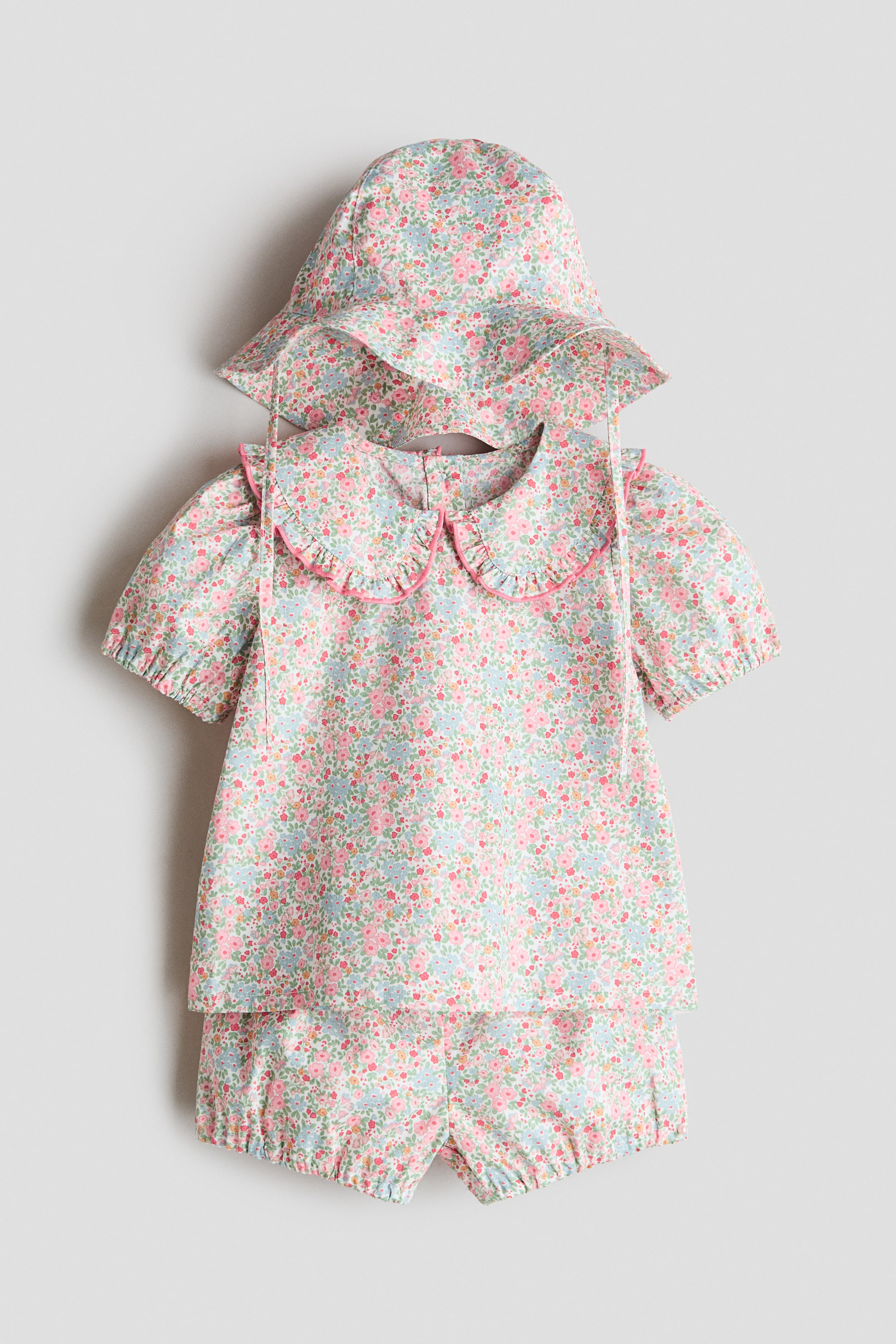 View larger image: A cotton seersucker set, showcased from the front, features a blouse, bloomers, and a sun hat, all adorned with a light blue and pink floral pattern. The blouse boasts a rounded, frill-trimmed collar with pink piping and puff sleeves with elastic cuffs, while the bloomers offer gentle elastic around the legs. The sun hat highlights a sloping brim with chin ties.