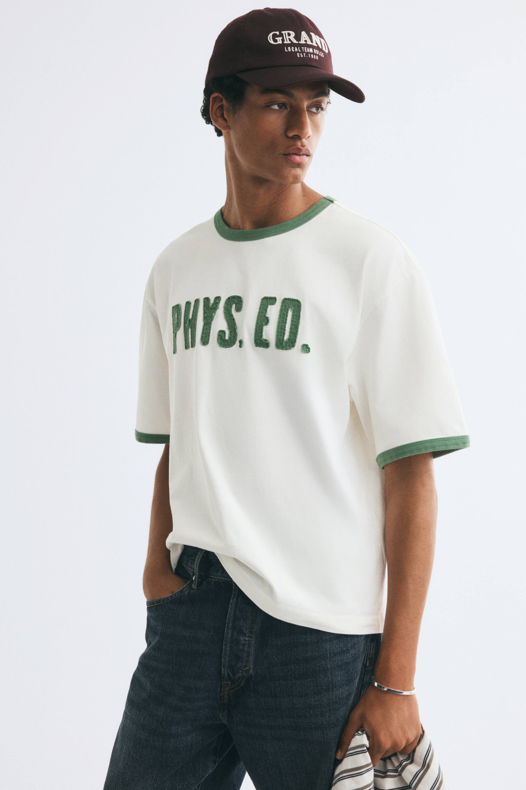 Vezi imaginea mai mare: Un tricou alb tip ringer, cu inscripția „PHYS. ED.” texturată în verde pe piept și cu margini contrastante verzi la guler și la manșetele mânecilor, este purtat de model. Modelul asortează tricoul cu blugi bleumarin închis și o șapcă de baseball maro.