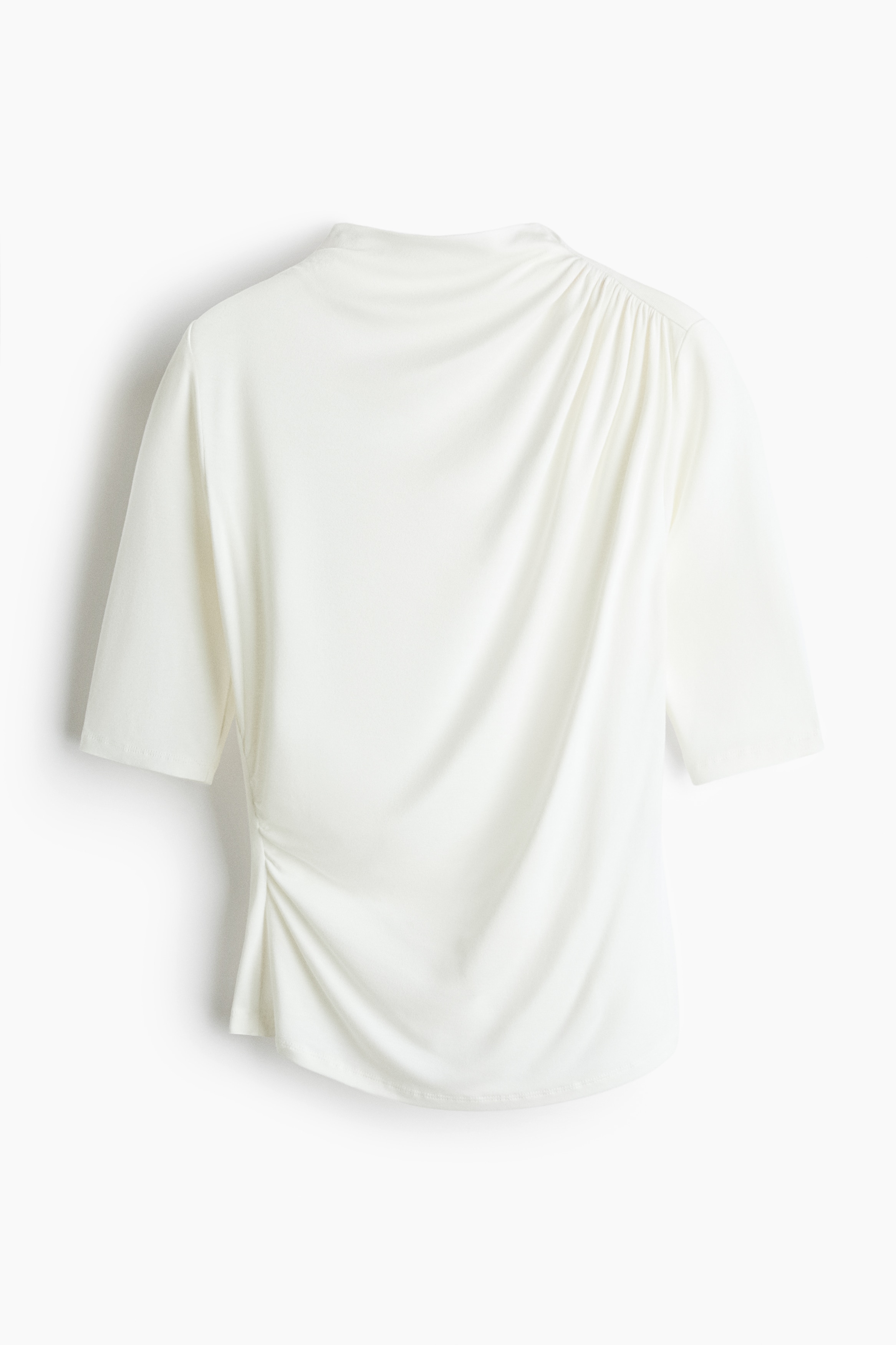 La modelo lleva una blusa blanca ajustada con cuello alto, mangas a la altura del codo y un detalle fruncido que crea un efecto drapeado en la parte delantera. La blusa se combina con pantalones blancos de tiro bajo.
