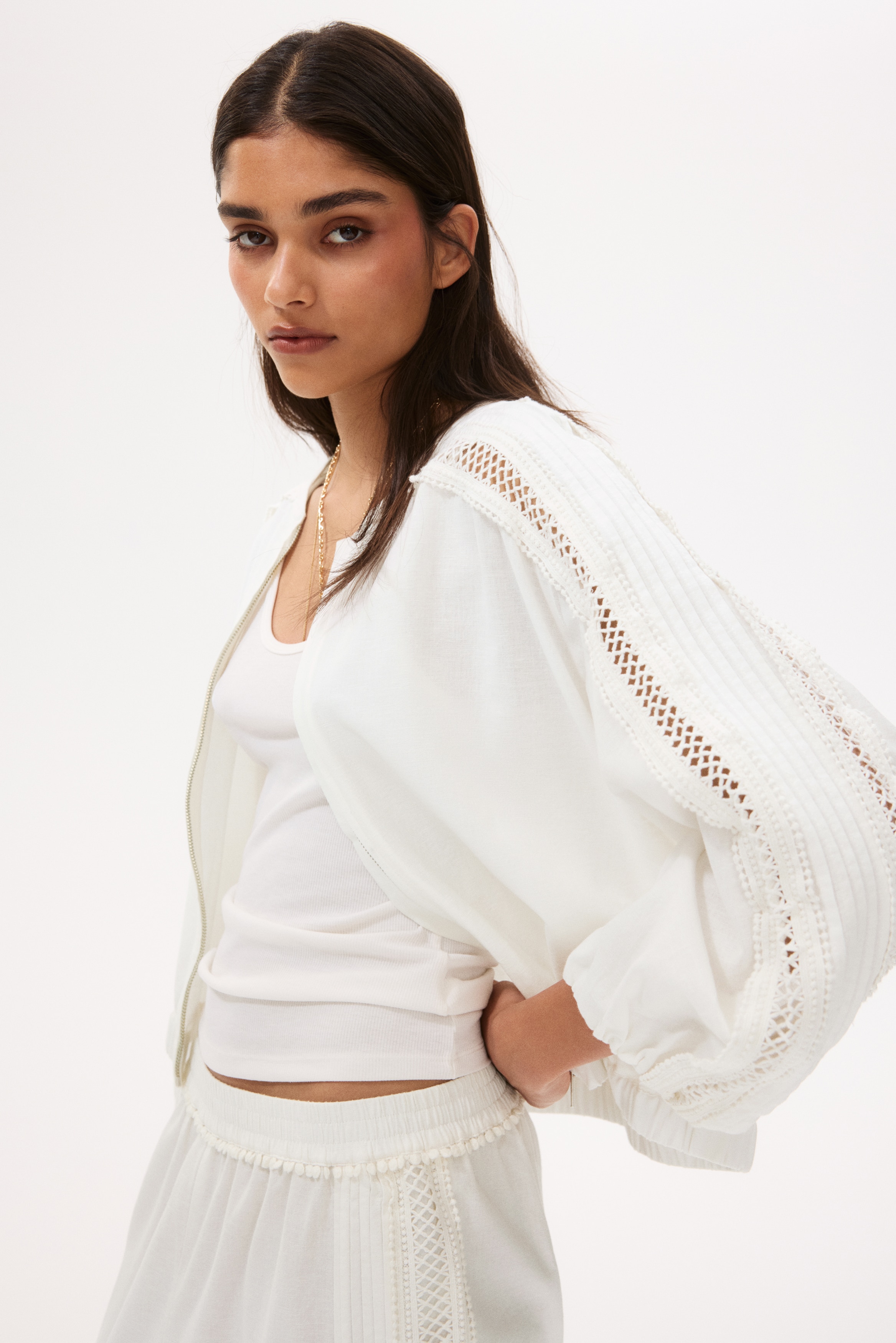 "A white bomber jacket is displayed prominently, highlighting its front with a full-zip closure and a rounded, collarless neckline. The raglan sleeves are adorned with intricate lace panels along the shoulders and arms, and both the sleeve cuffs and bottom hem are elasticated."