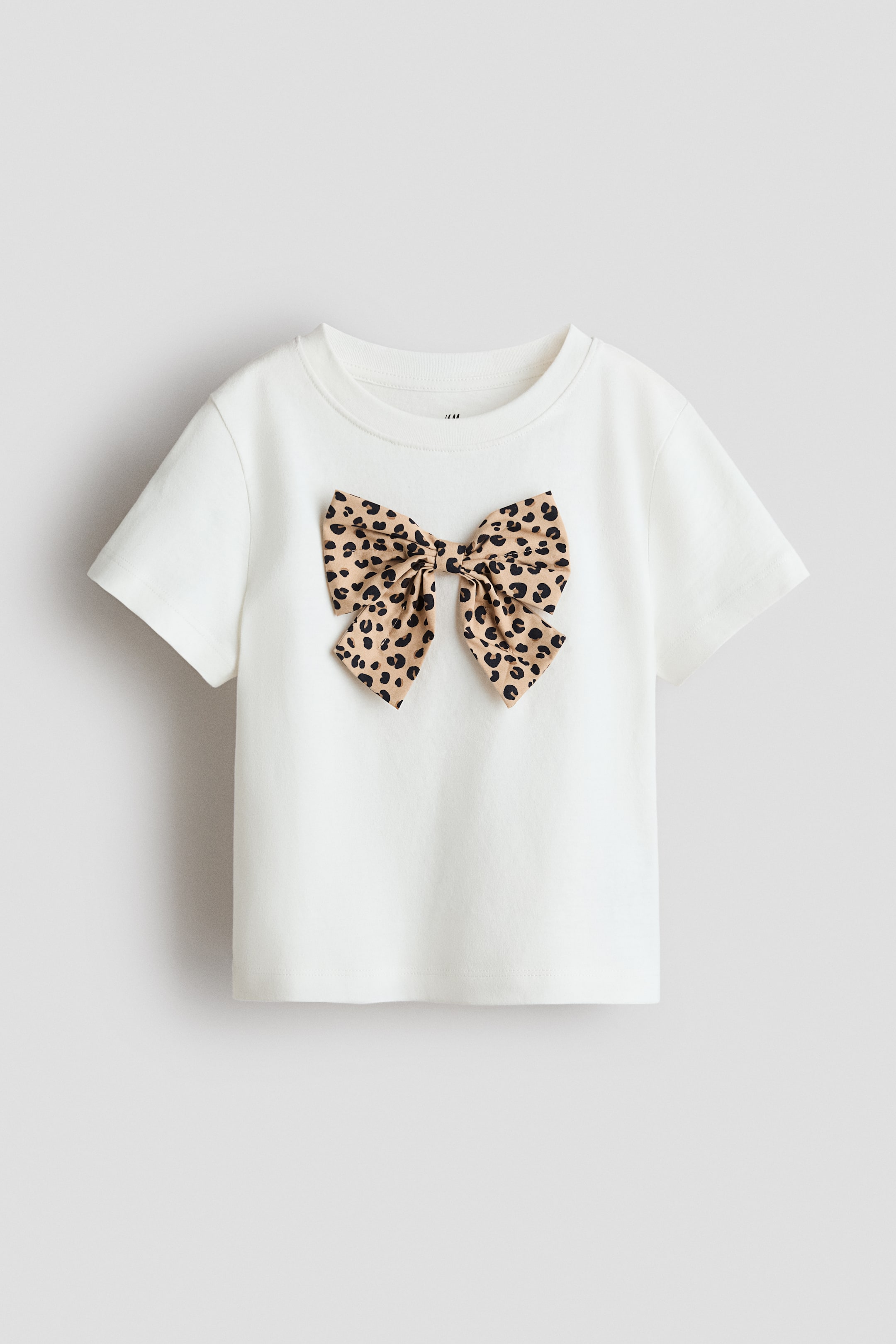 הצגת תמונה גדולה יותר: טי-שירט בצבע אוף-וייט מעוטרת בפפיון גדול מקופל בדוגמת נמר בחזיתה. היא מעוצבת עם צווארון עגול בגימור ריב ושרוולים קצרים.