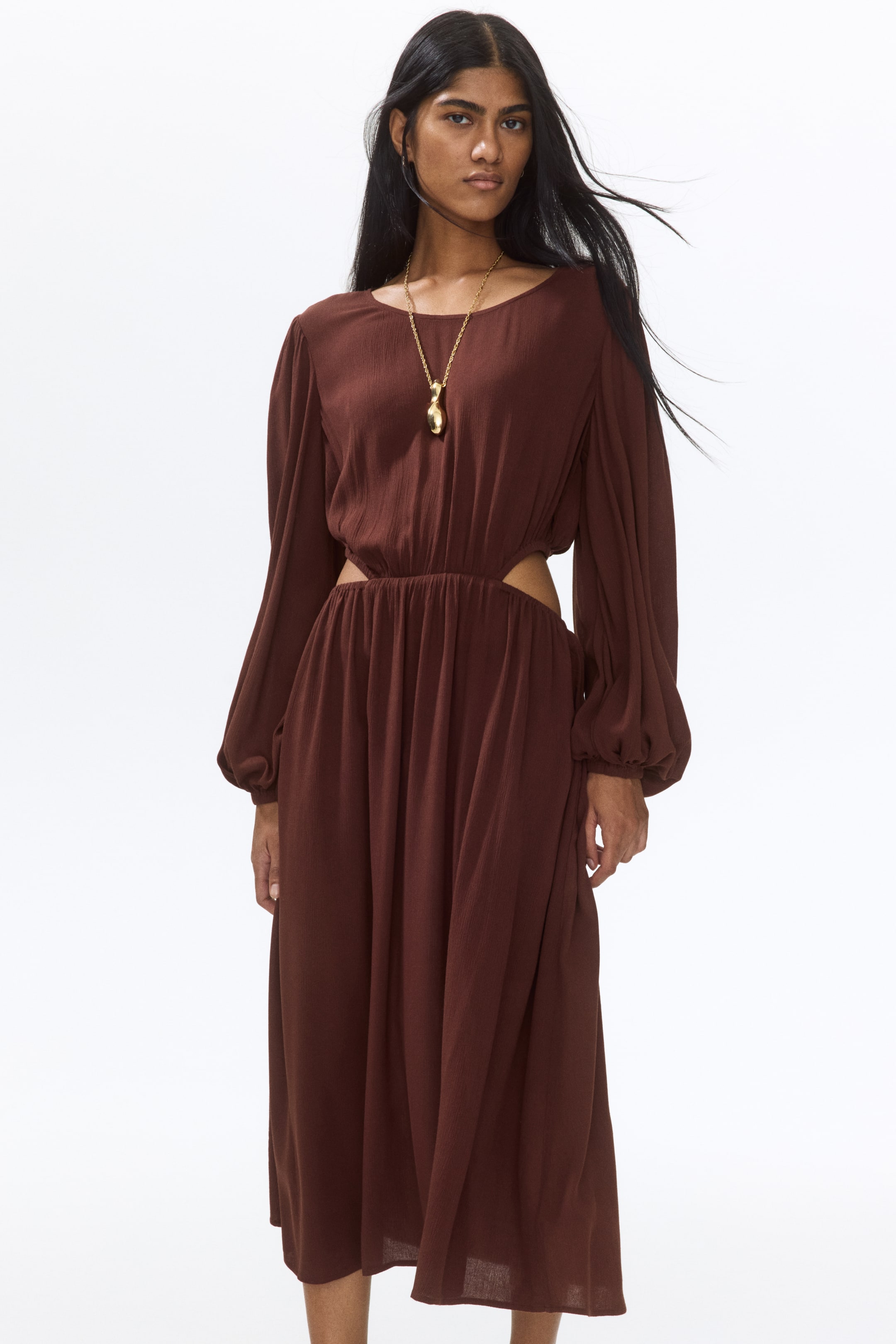 View larger image: The model is adorned in a dark brown calf-length dress, featuring a round neckline and long balloon sleeves. The dress boasts a gathered waist with cut-out sections on the sides and a gently flared skirt. Complementing the ensemble, the model dons a gold-toned necklace.