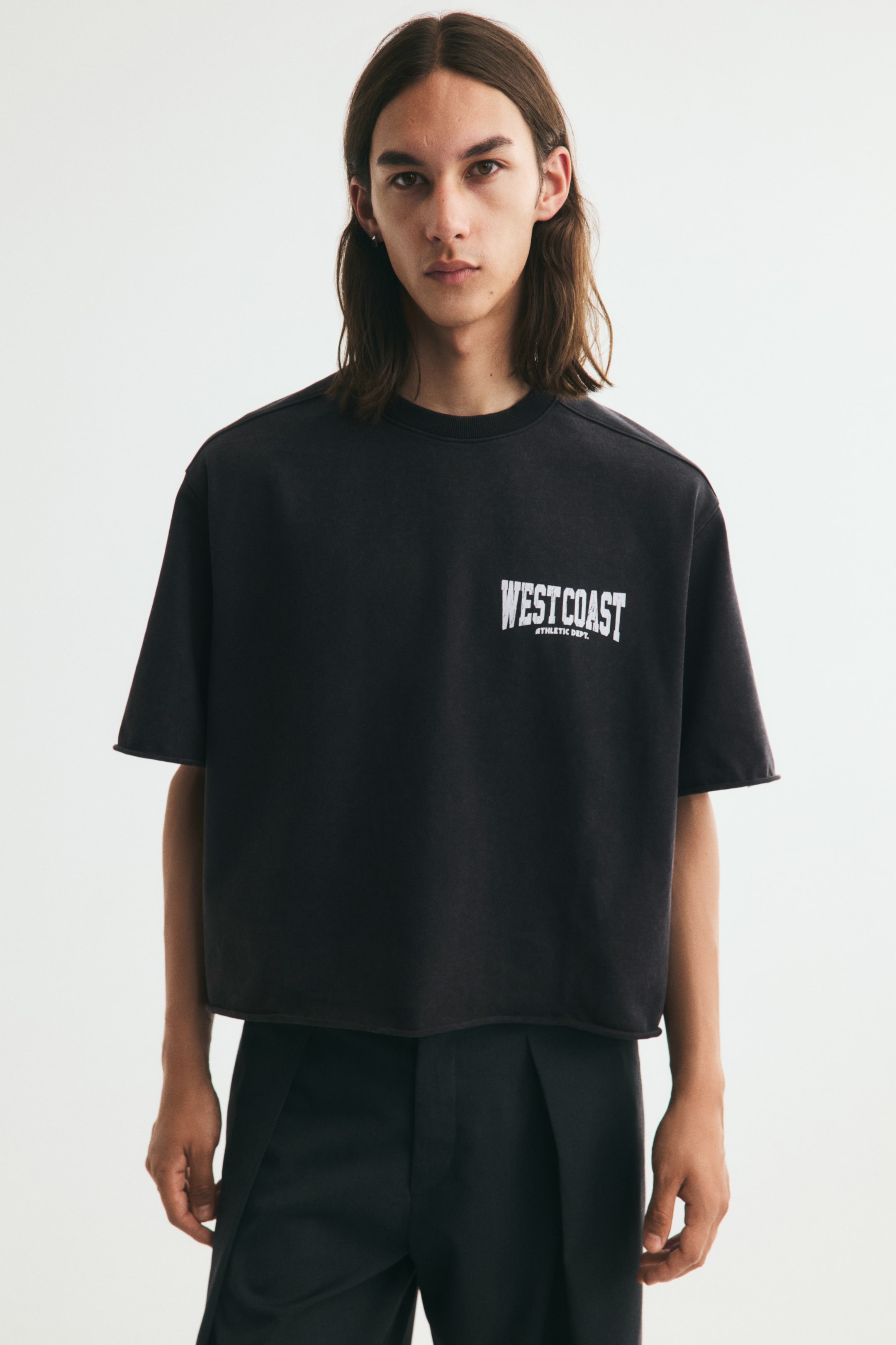 View larger image: A short, boxy black T-shirt with an oversized fit is worn by the model, paired with dark trousers. The T-shirt features dropped shoulders, a round ribbed neckline, raw-cut edges at the cuffs and hem, and a white text print reading "WEST COAST" on the left chest.