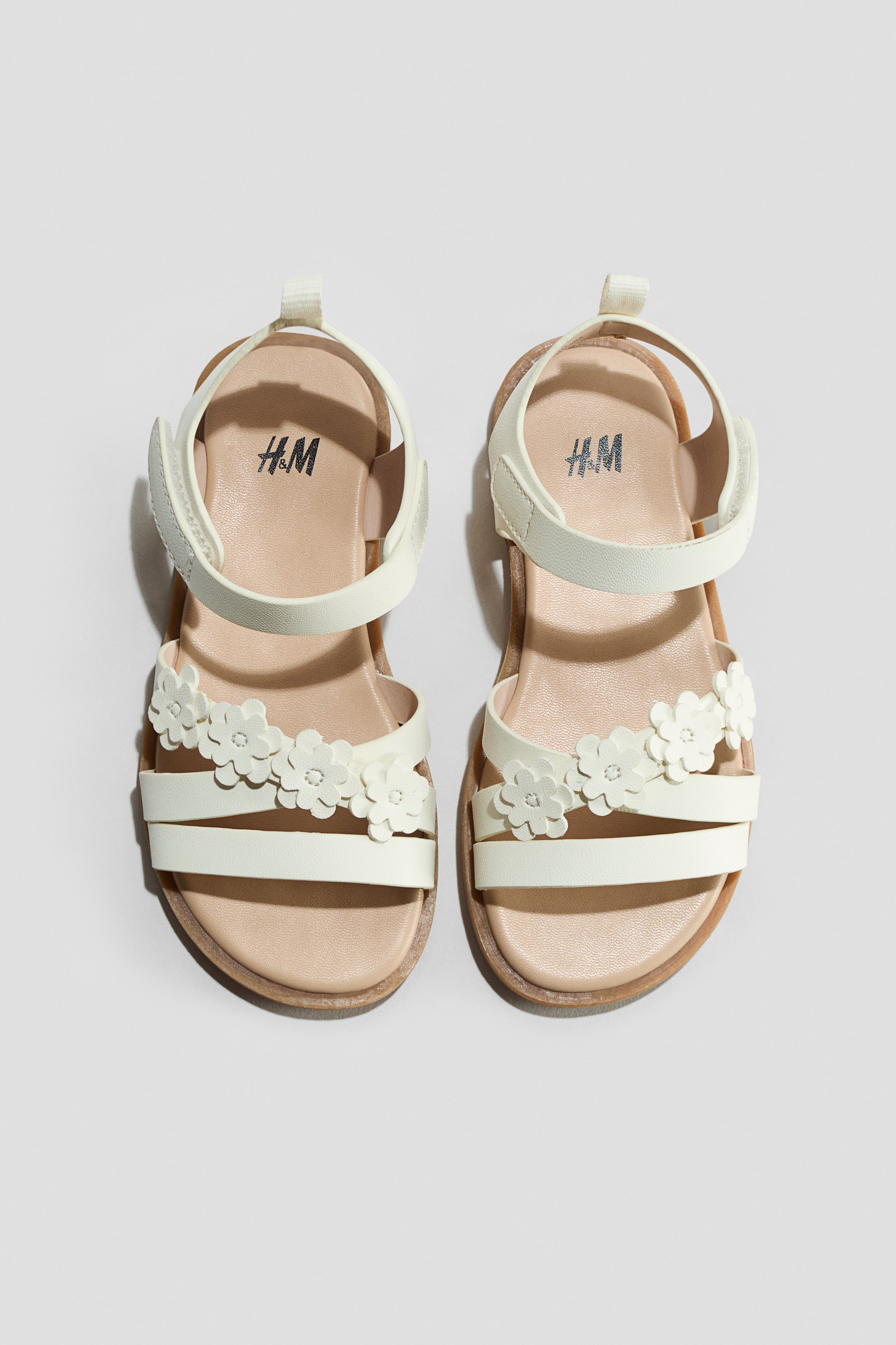 View larger image: A pair of cream sandals is shown from the front, featuring criss-cross straps over the foot adorned with flower appliqués. An adjustable hook-and-loop fastening secures each sandal around the ankle, with the H&M logo visible on the insole.