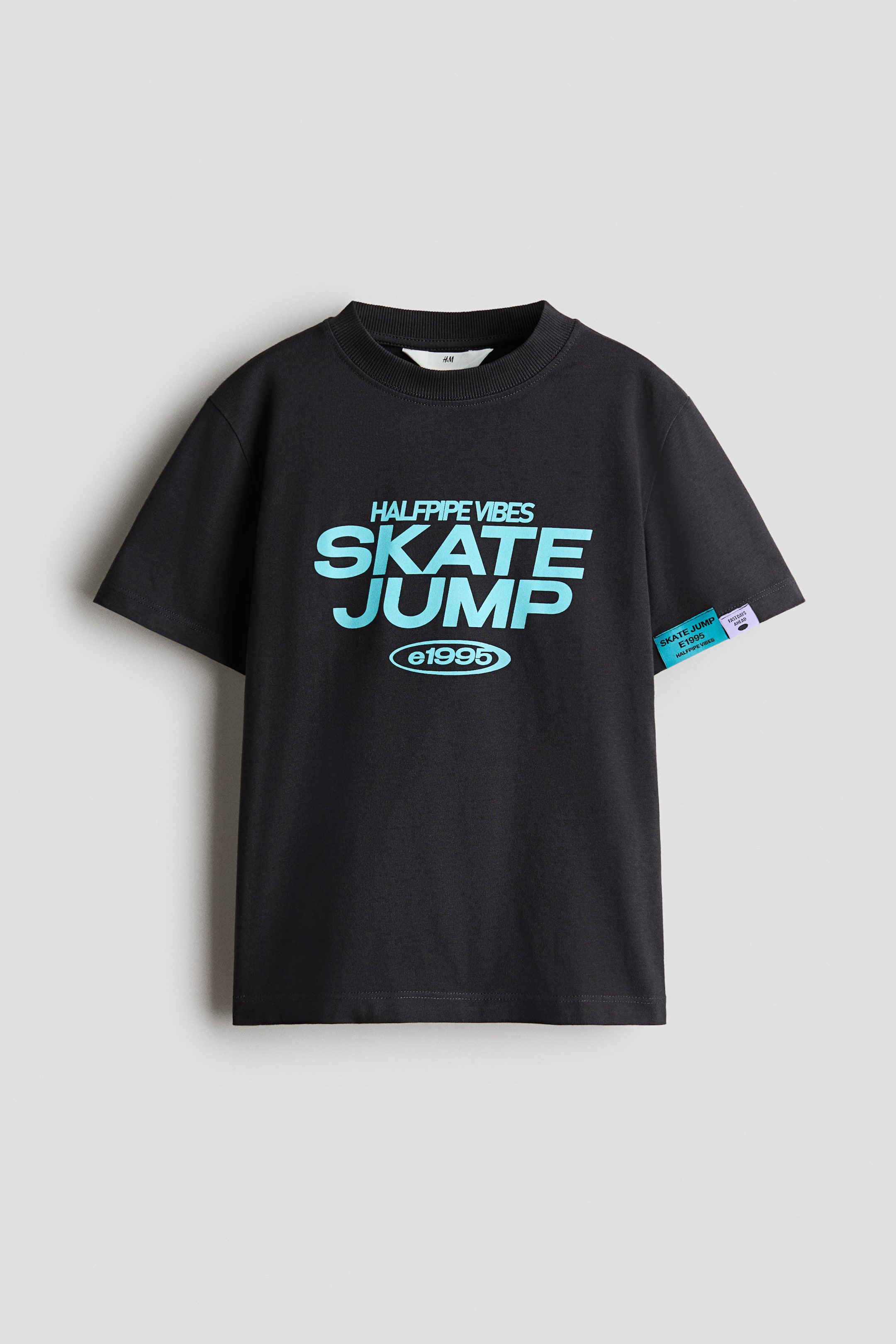 View larger image: A black t-shirt is laid flat, showcasing its front with a round, ribbed neckline and dropped shoulders. A blue and teal gradient graphic print reading 'HALFPIPE VIBES SKATE JUMP ©1995' is prominently displayed in the center, and a blue fabric tag with text is visible on the right sleeve.