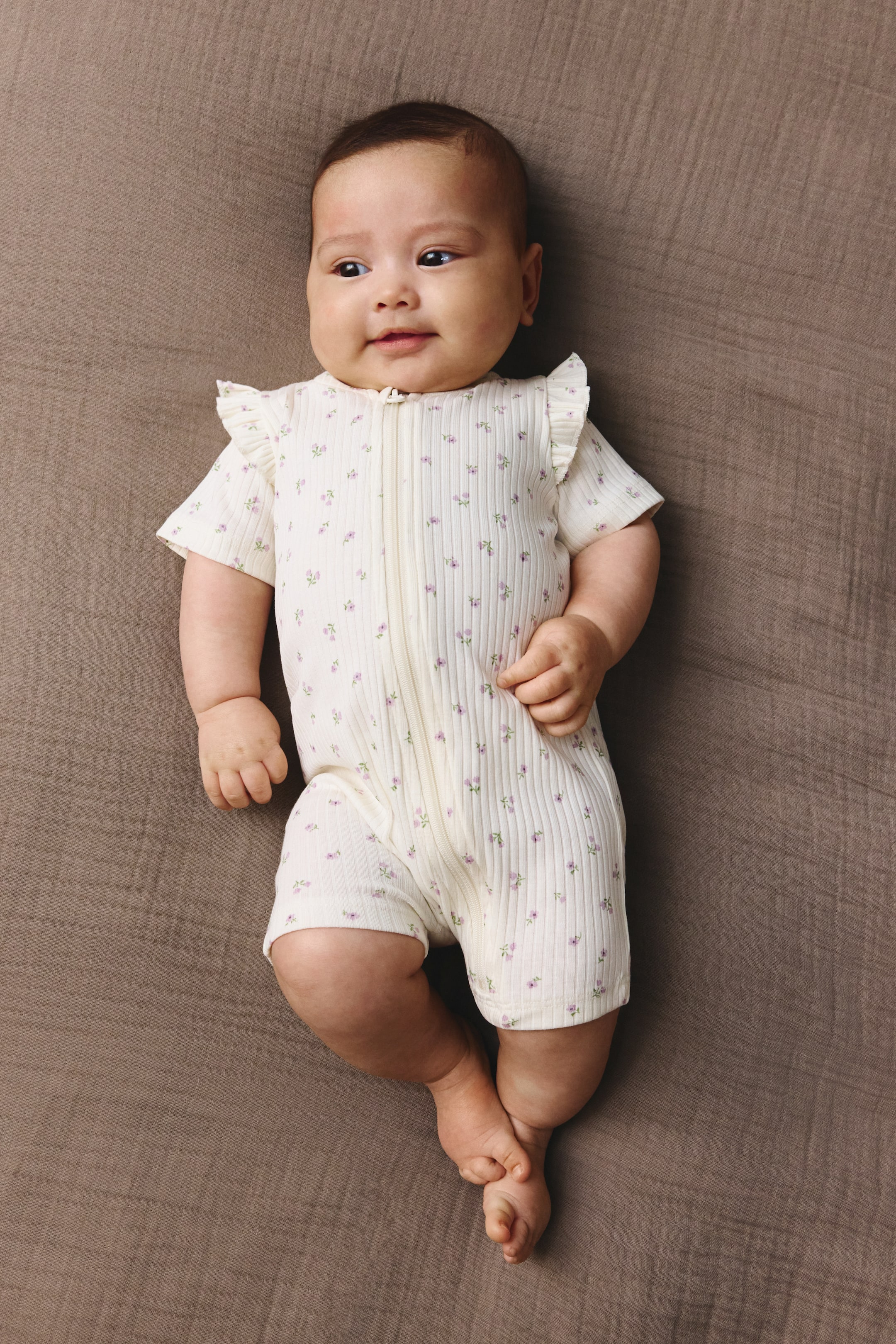 View larger image: The cream ribbed knit baby romper features short sleeves with ruffled shoulders and a full-length front zipper. It's adorned with small purple floral designs and ends just above the knee.
