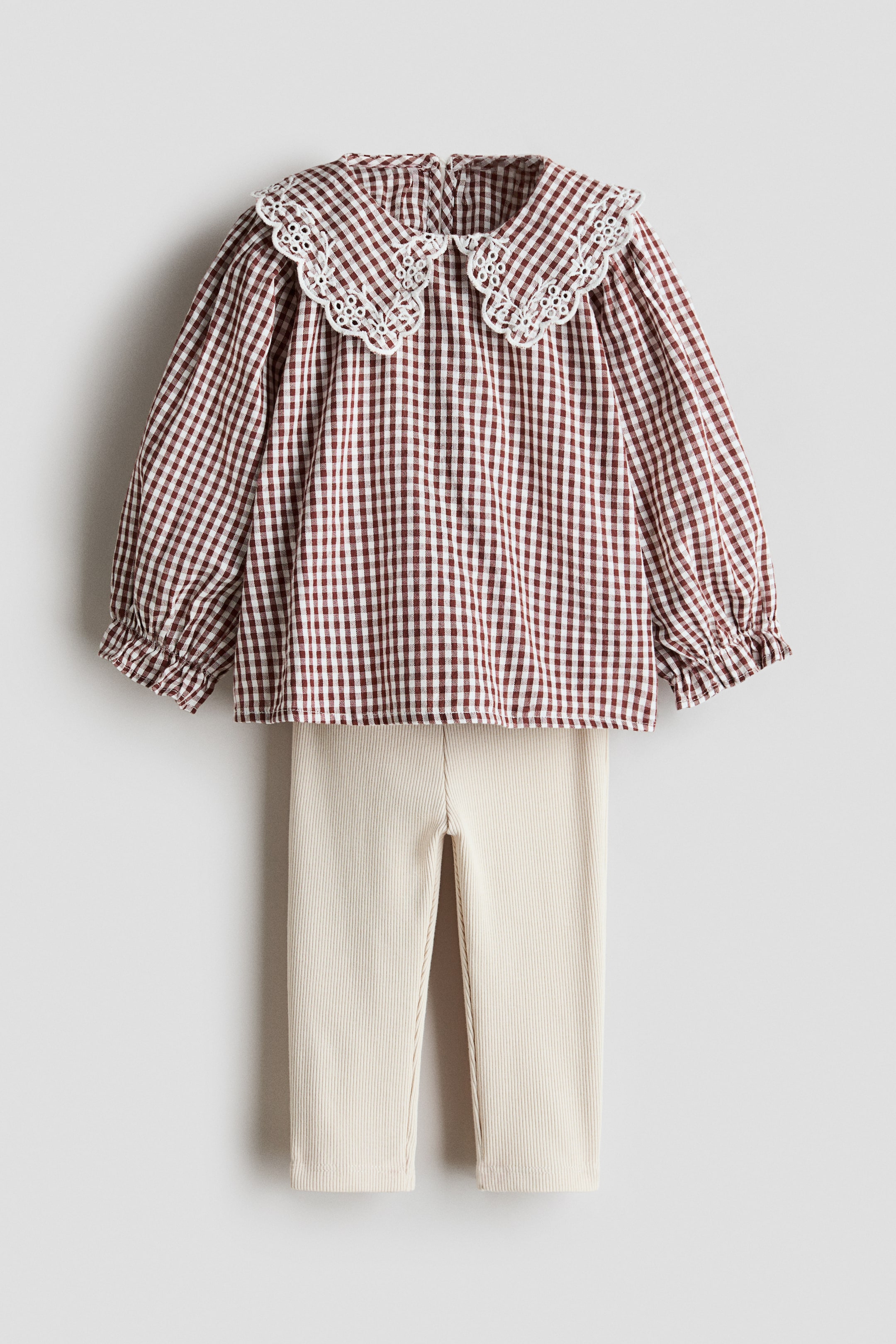 View larger image: A two-piece outfit features a brown and white checkered blouse with a large white scalloped eyelet lace collar and puffed sleeves with elasticated cuffs. It is paired with ribbed, light beige straight-leg trousers.
