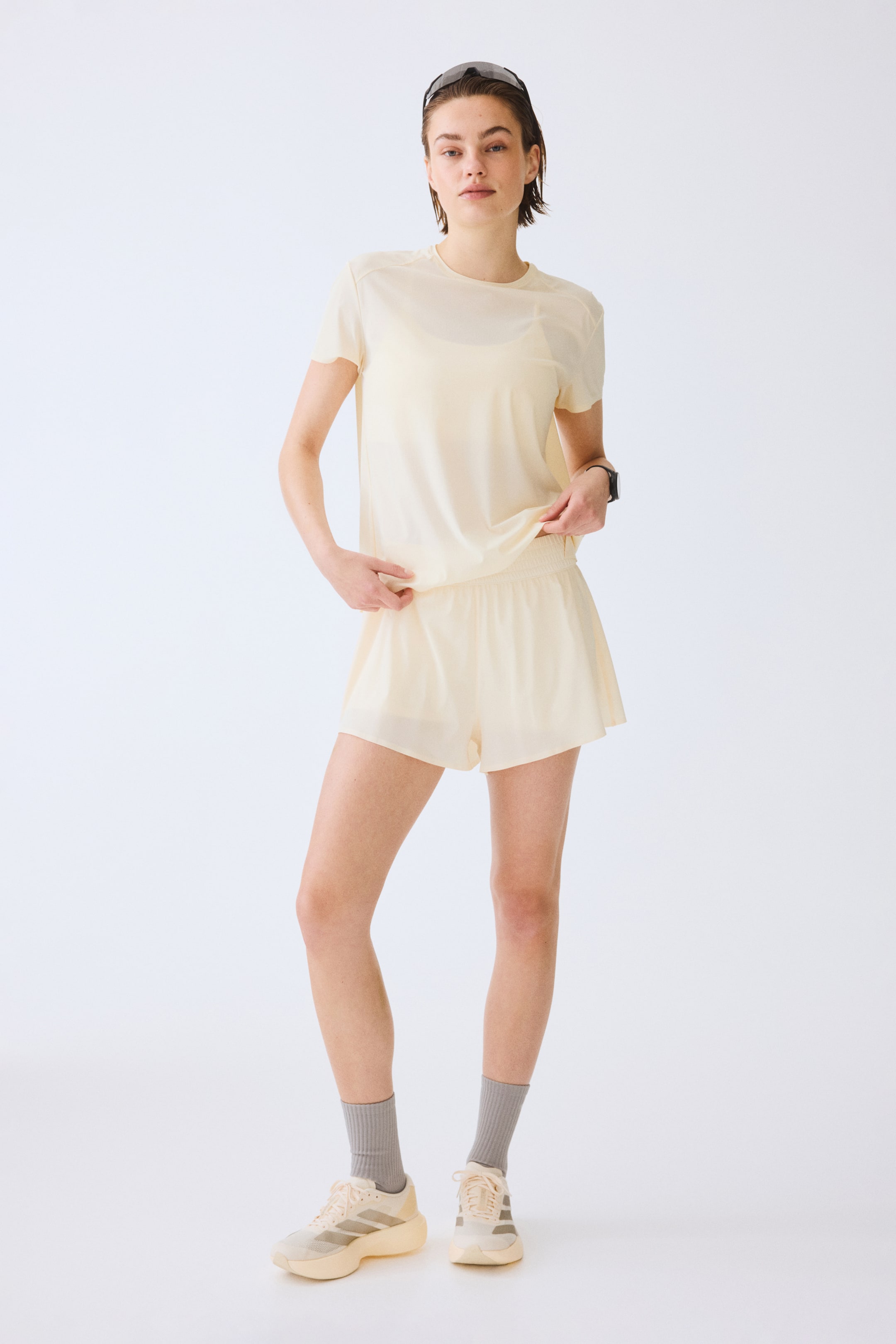 Zobraziť väčší obrázok: Svetložlté krátkorukávové športové tričko je prezentované spredu, vrstvené na zodpovedajúcej športovej podprsenke a jemne zdvihnuté na leme, v kombinácii so svetložltými športovými šortkami. Tričko má raglánové rukávy a zaoblený výstrih, zatiaľ čo šortky majú elastický pás a voľný strih. Svetlé športové tenisky s bočnými pruhmi a sivé ponožky po členky dokončujú celkový vzhľad.