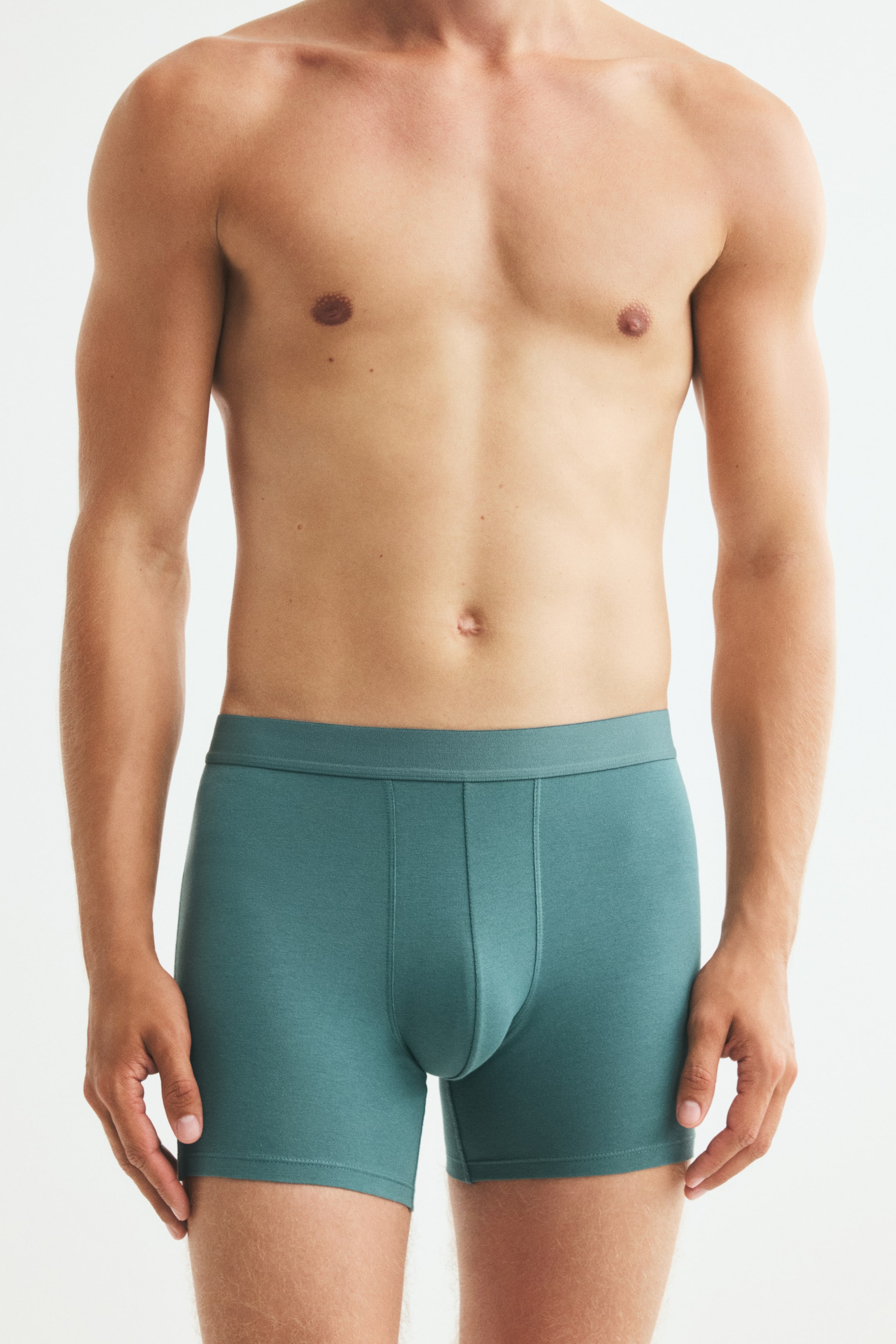 View larger image: The model is wearing teal boxer briefs, featuring a flat, elastic waistband and a contoured front pouch with vertical seams. The close-fitting underwear extends to the upper thighs.