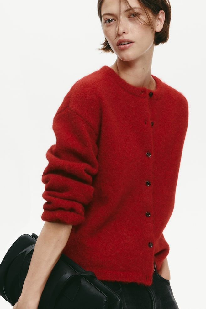 Ein roter Cardigan aus strukturiertem Strick mit Rundhalsausschnitt, Knopfleiste vorn und überschnittenen Schultern wird von dem Model getragen. Das Model kombiniert den Cardigan mit einer schwarzen Hose und hält eine schwarze Ledertasche unter einem Arm. - 1