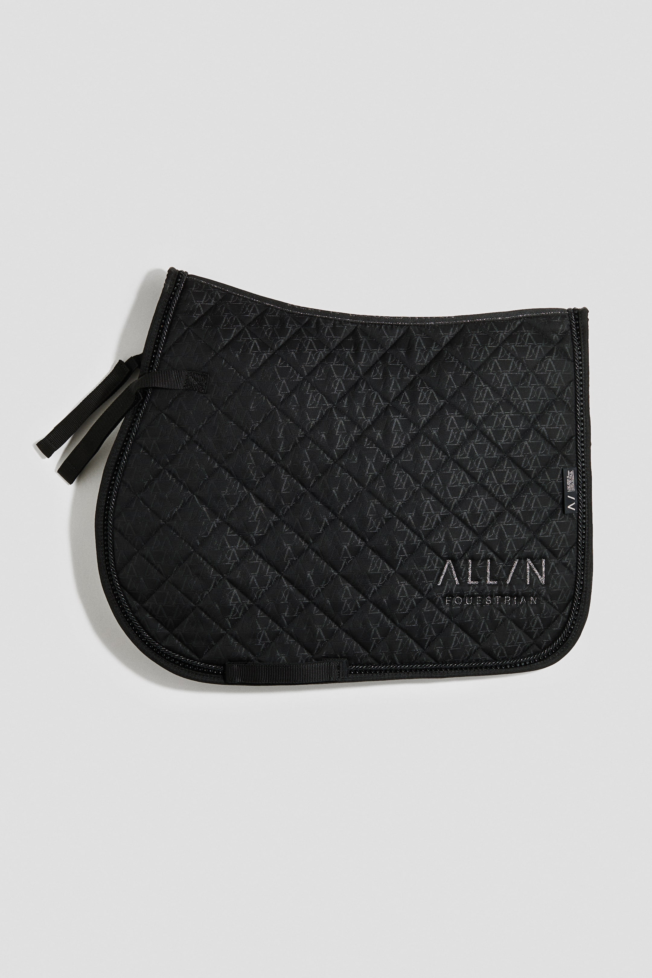查看大圖: 一個黑色的馬鞍墊呈現出帶有微妙幾何圖案的絎縫表面，菱形縫線中可見其圖案。它的邊緣都配有黑色編織的滾邊、多條黑色布帶用於固定，右下角還有銀色文字的「ALL/N EQUESTRIAN」標誌。