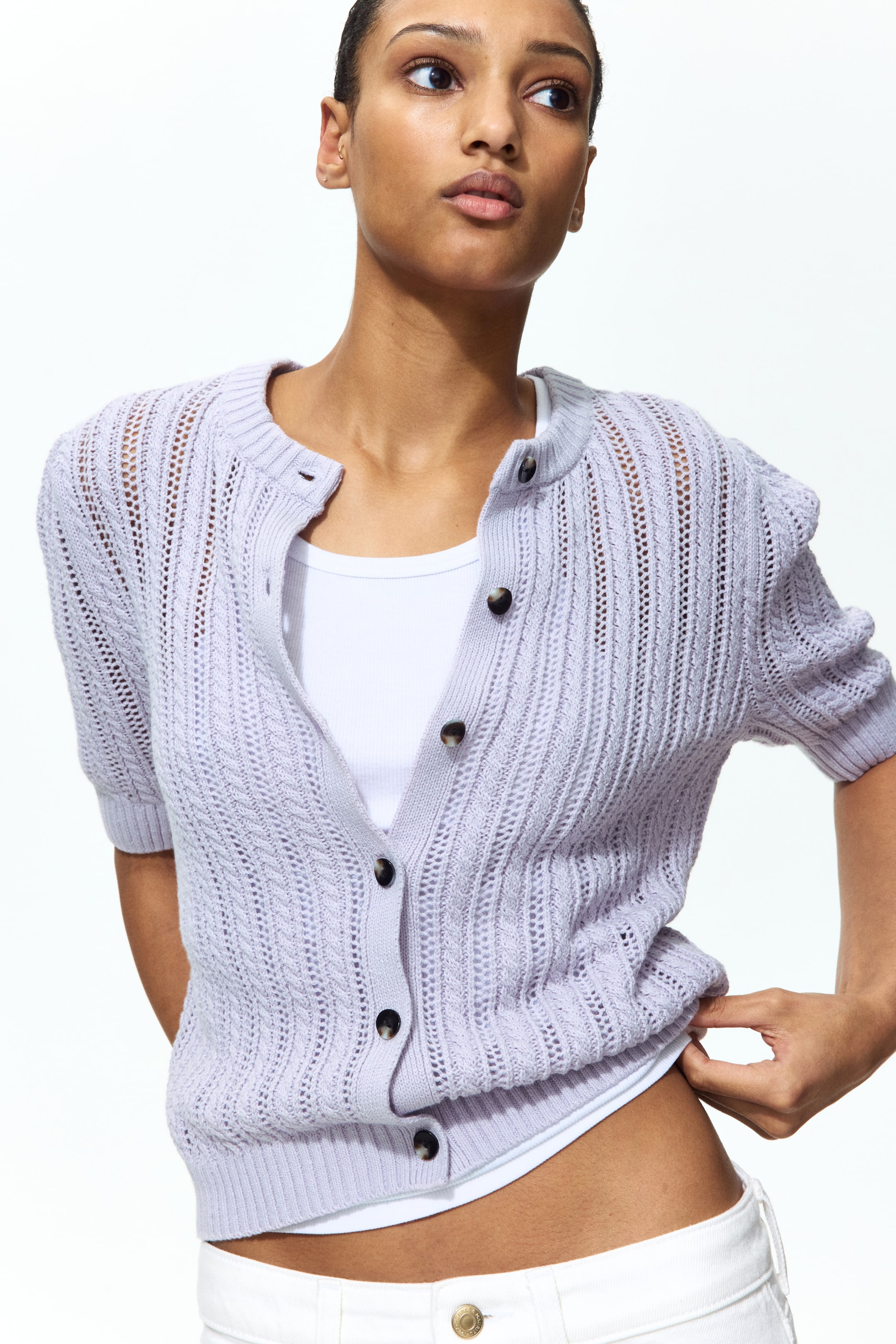 View larger image: A light purple short-sleeve cardigan is shown from a front-quarter angle, featuring a round neck and an open-knit pattern with vertical detailing. It is partially unbuttoned down the front with dark buttons, worn over a white tank top, and showcases a ribbed hem.