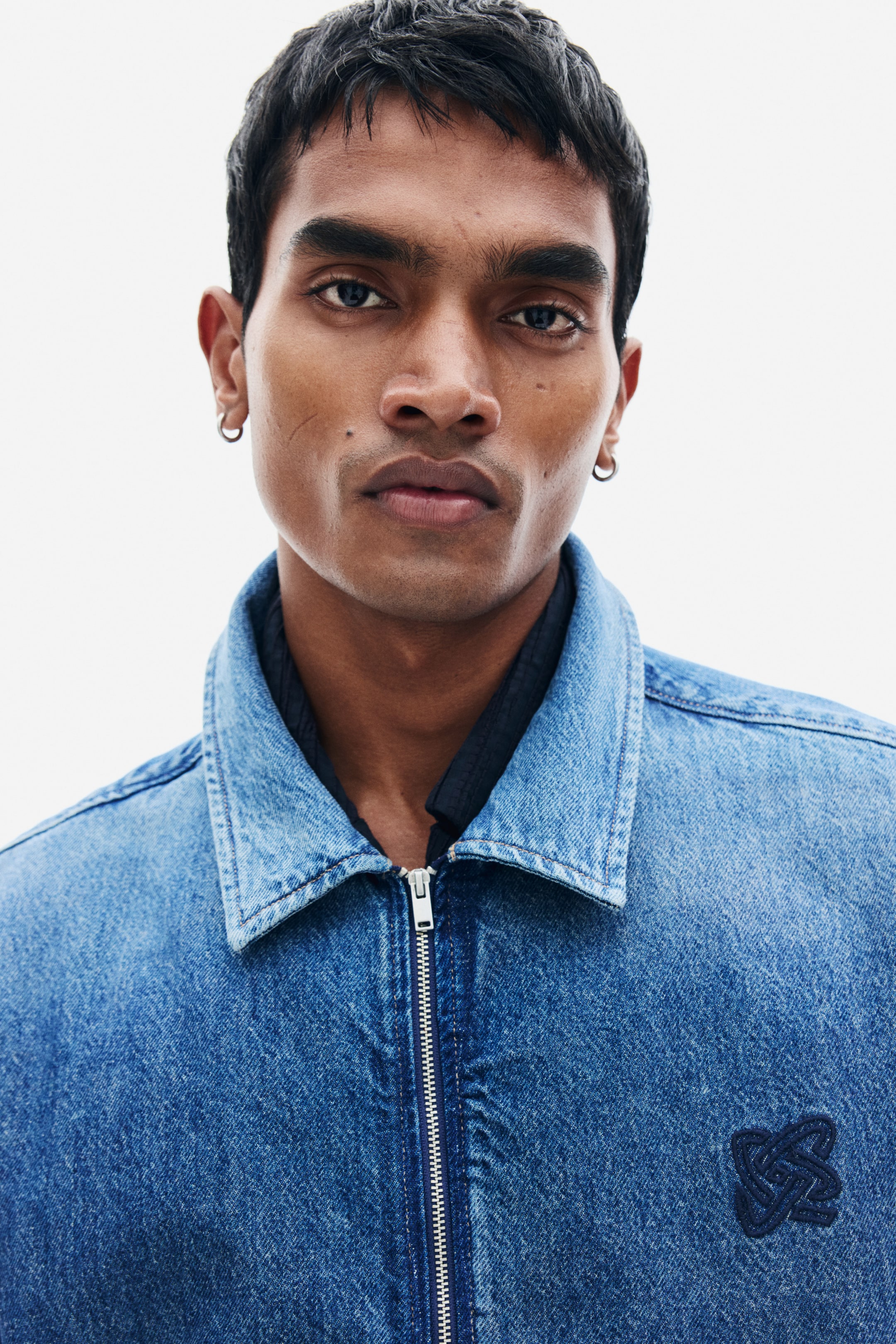 View larger image: A blue denim jacket featuring a front zip fastening, a classic pointed collar, and an embroidered logo on the left chest is worn by the model. The jacket is layered over a dark collared shirt.