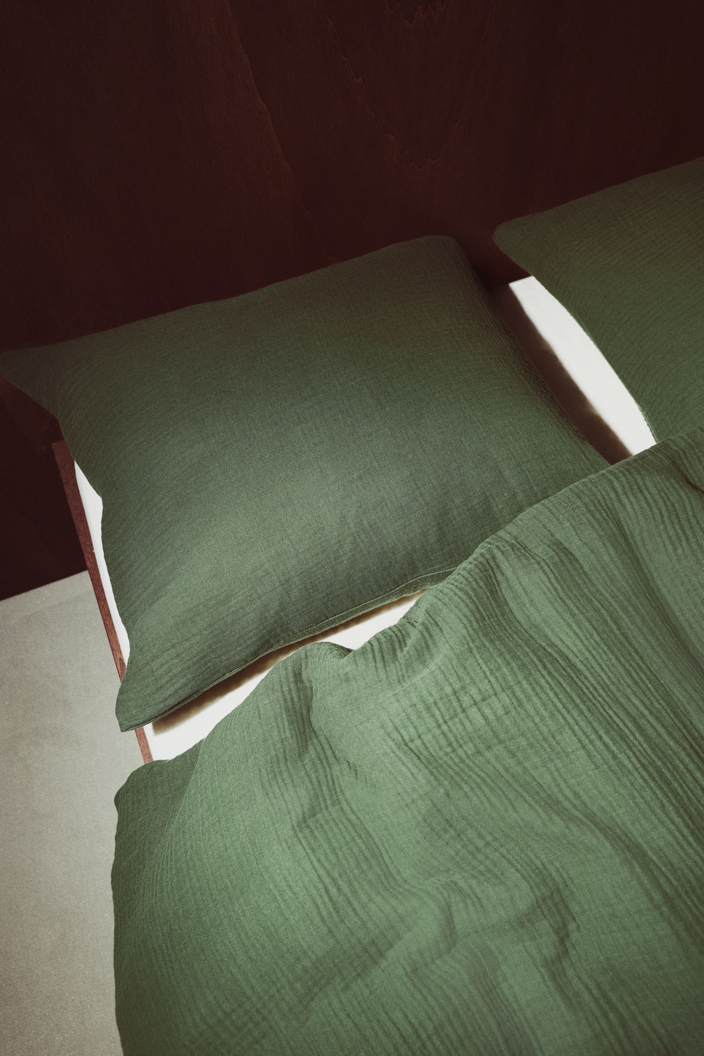 Un set di copripiumino in mussola di cotone verde scuro Ã¨ mostrato frontalmente, con il copripiumino ripiegato in modo ordinato appoggiato nella parte inferiore e una federa abbinata in posizione verticale dietro di esso. Entrambi i pezzi presentano un aspetto testurizzato e stropicciato.